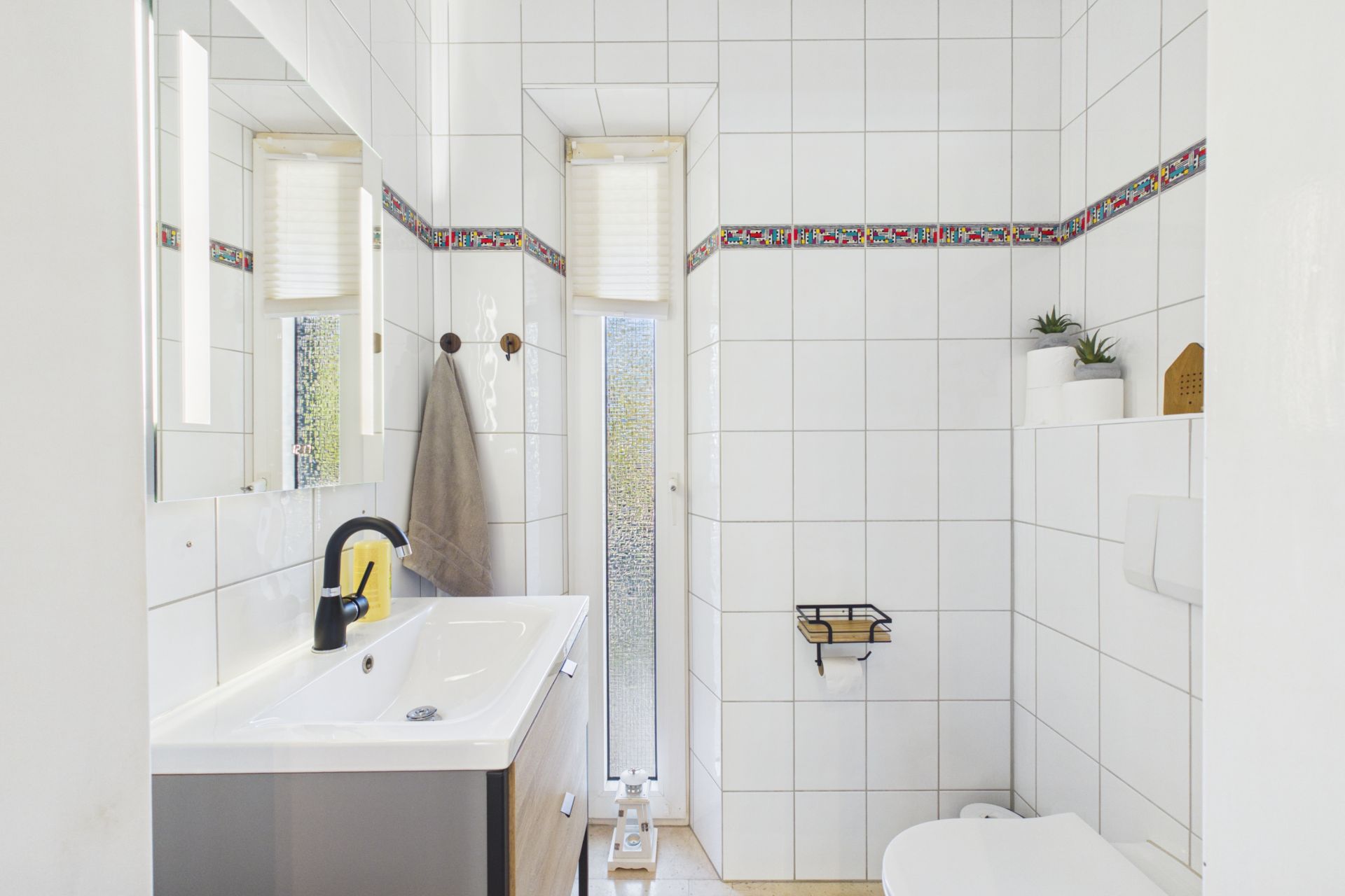 Helles Badezimmer mit weiß gefliesten Wänden, Waschbecken, Spiegel, Toilette und kleinen Pflanzen.