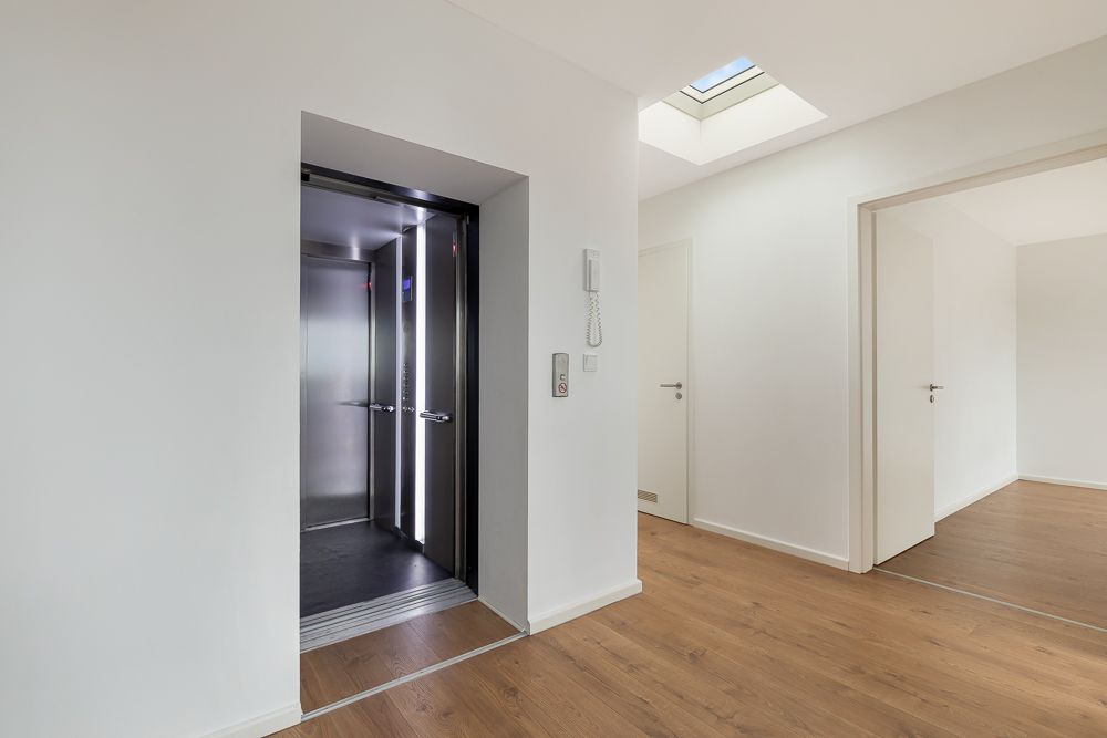 Heller Flur mit offenem Aufzug, Holzboden und mehreren weißen Türen.