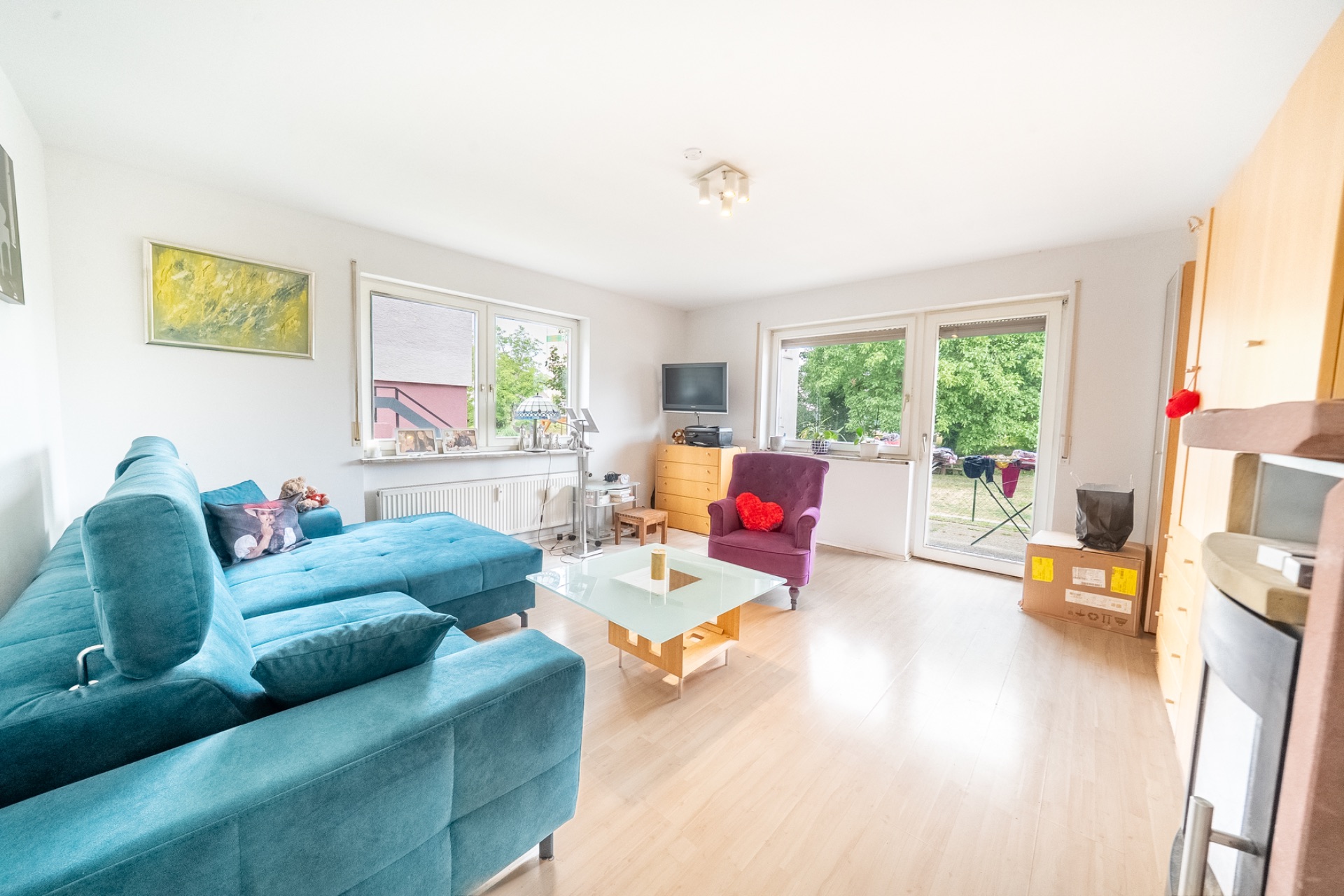 Helles Wohnzimmer mit blauem Sofa, Glastisch, Sessel und großem Fenster mit Blick ins Grüne.