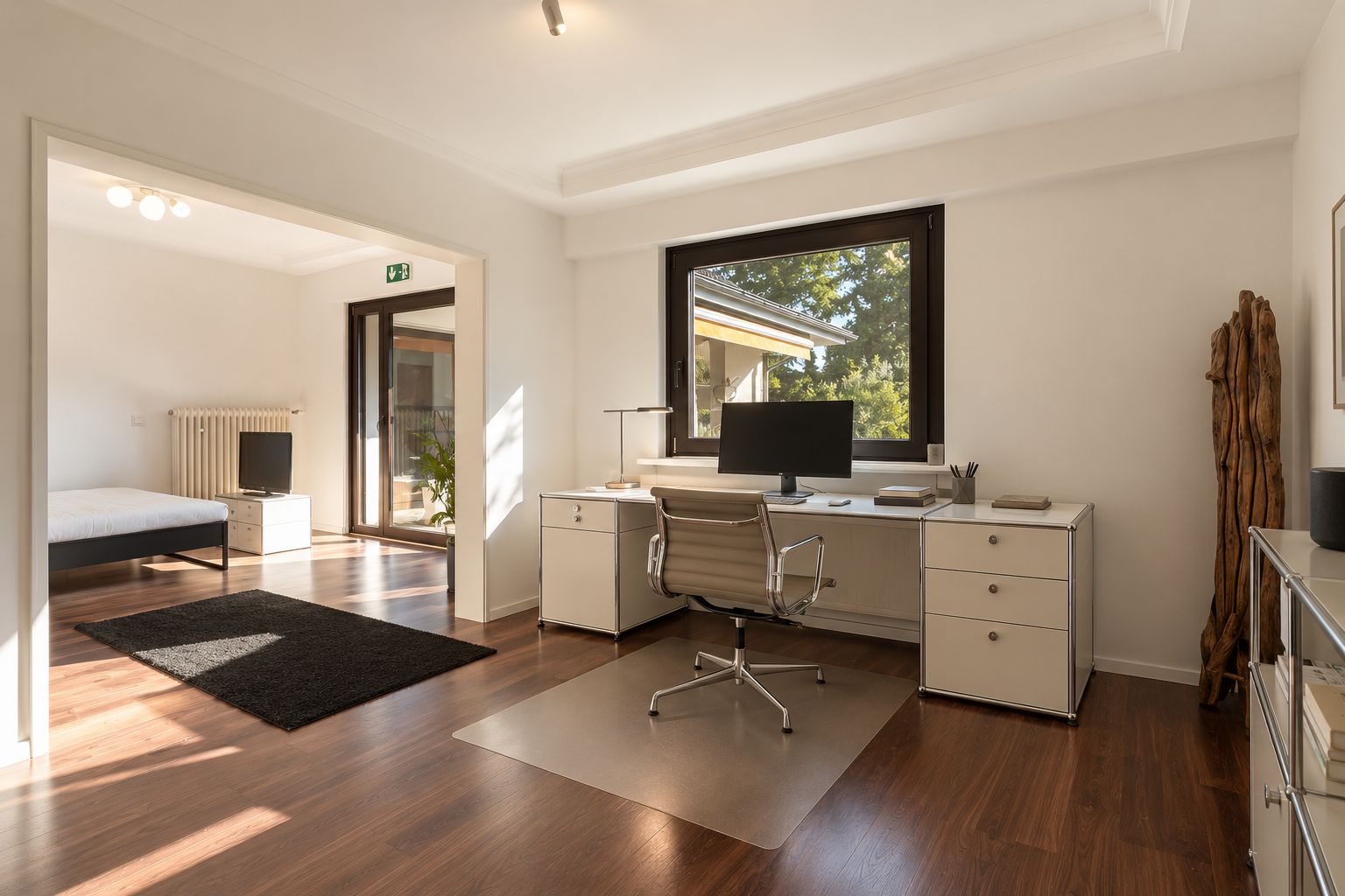 Helles Arbeitszimmer mit Schreibtisch, Stuhl, Computer und großem Fenster, daneben ein offenes Schlafzimmer.