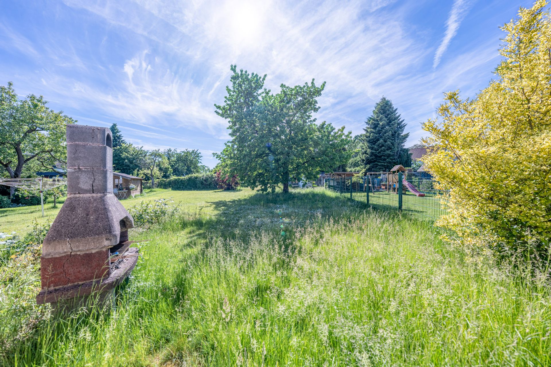 Großer Garten mit gemauertem Grill, Bäumen und Rasen unter blauem Himmel.