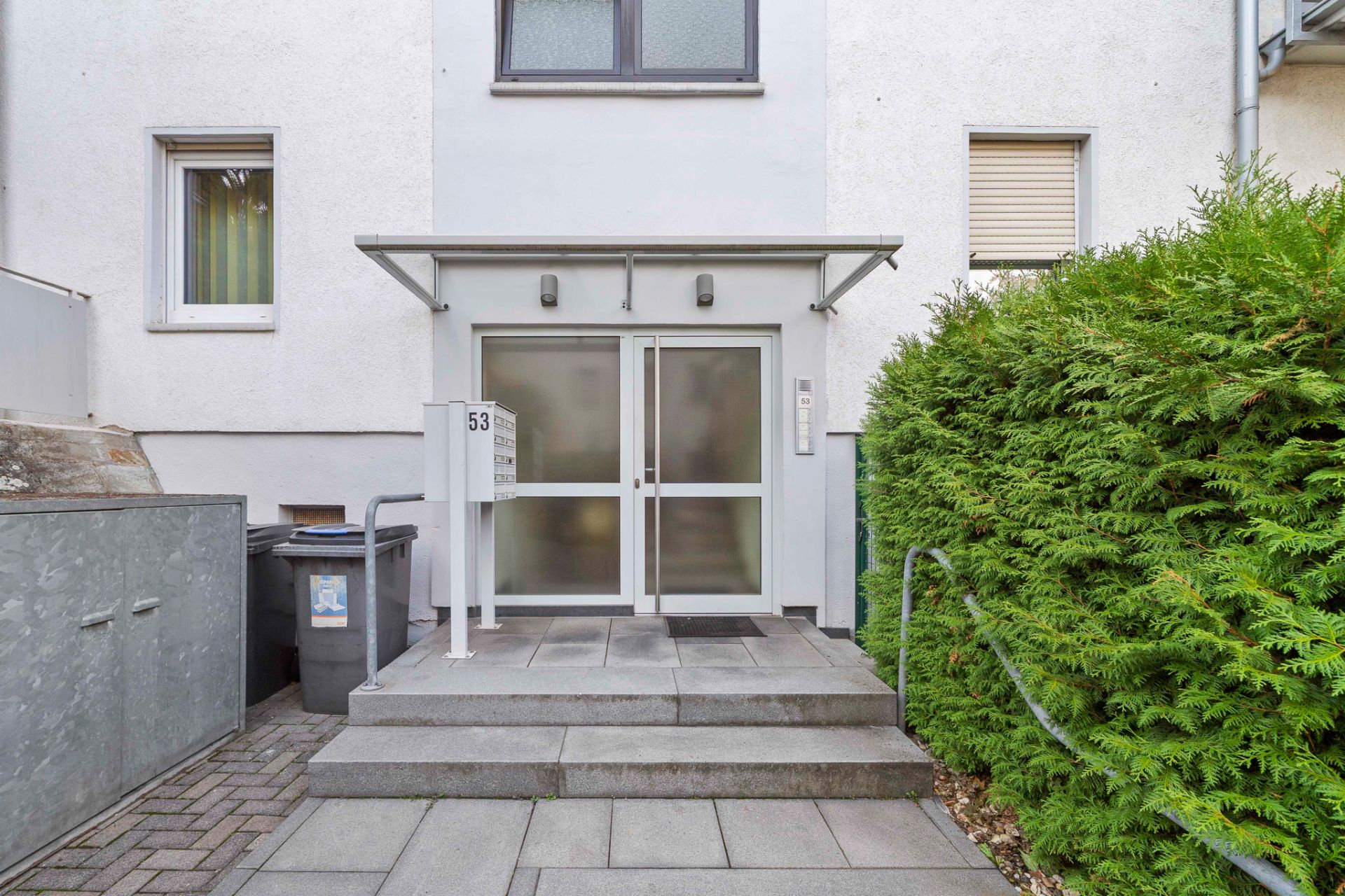 Wohnhauseingang mit Glastür, Briefkästen und Treppe, daneben Mülltonnen und grüne Hecke.