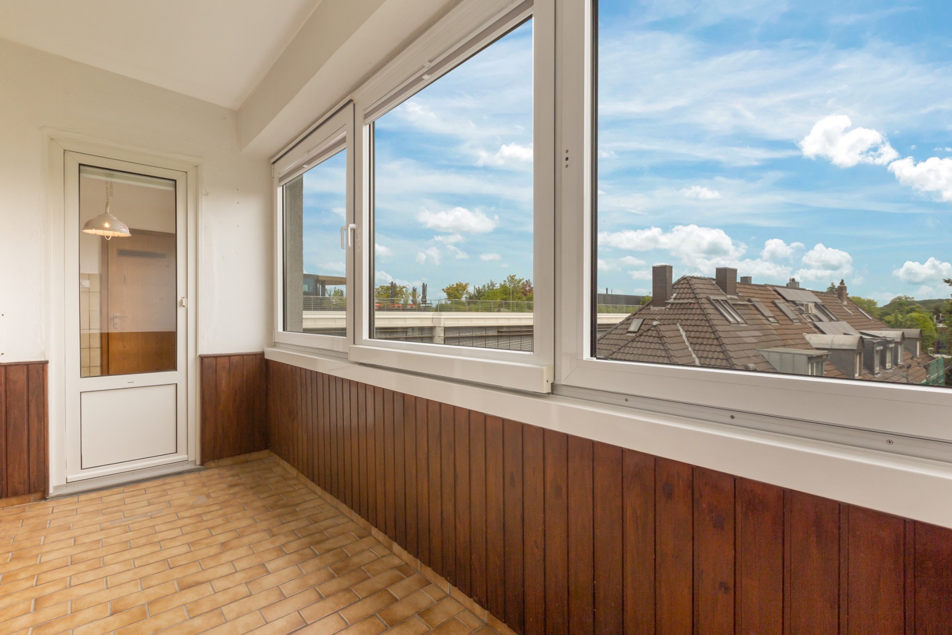 Geschlossene Loggia mit großen Fenstern, braunen Fliesen und Holzverkleidung.