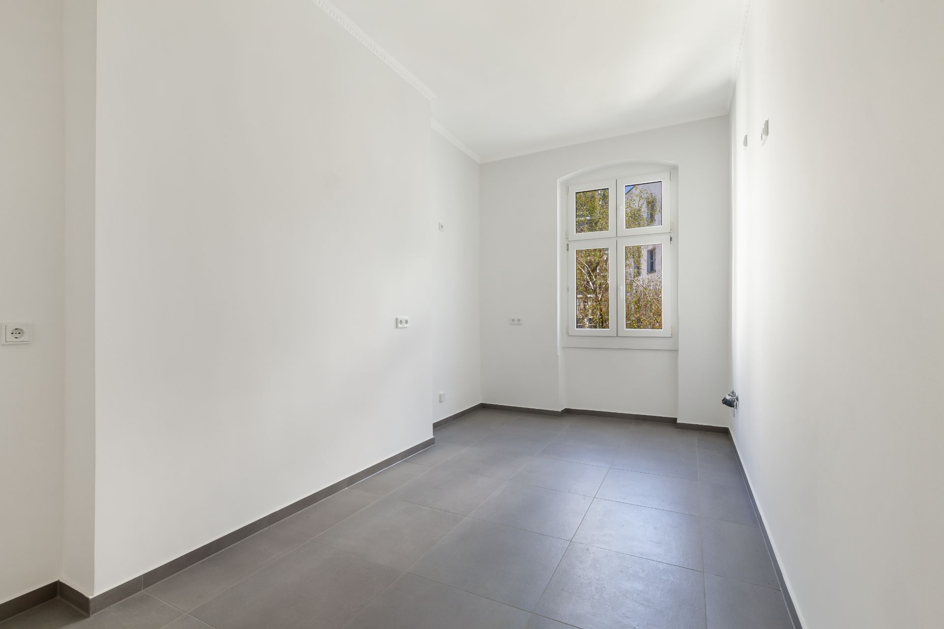 Leerer, heller Raum mit grauem Fliesenboden und großem Fenster.