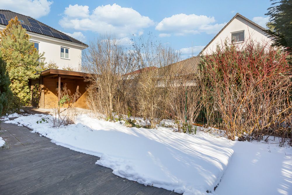 Verschneiter Garten mit Holzterrasse, Büschen und Haus im Hintergrund.