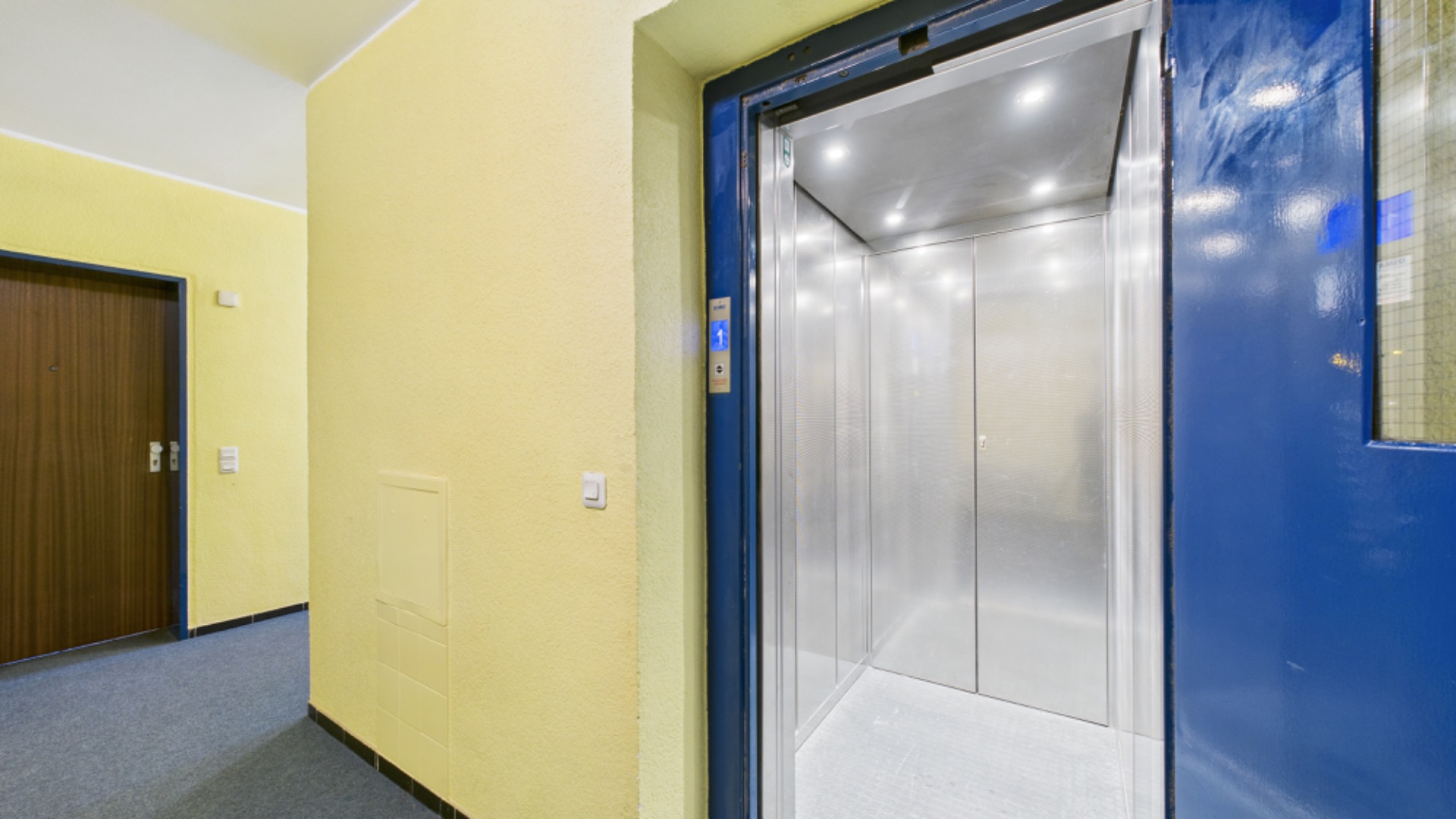 Geöffneter Aufzug neben Wohnungstür in einem hellen Flur mit gelben Wänden.