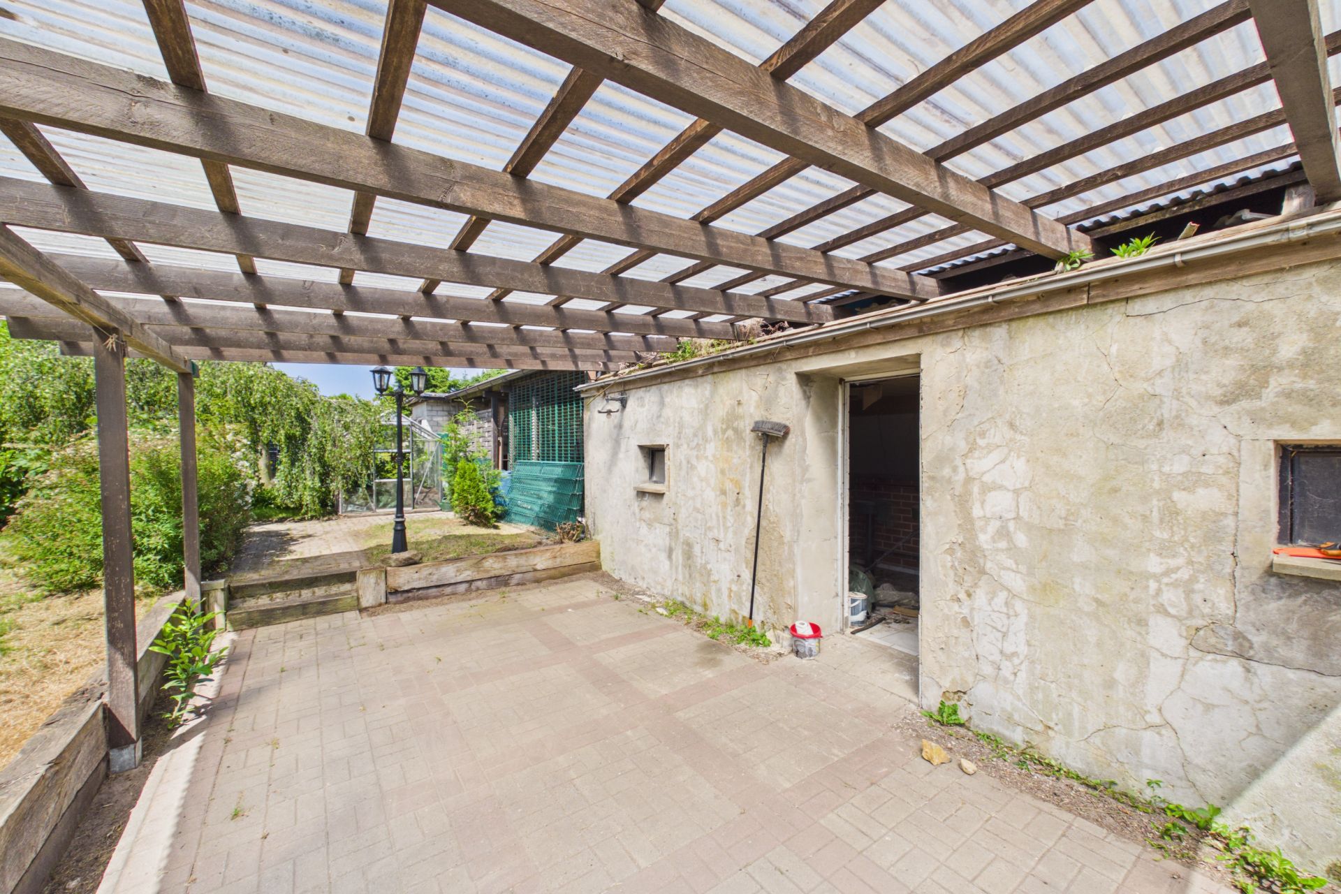 Überdachte Terrasse mit Pflastersteinen neben einem Haus mit rauer Steinwand.