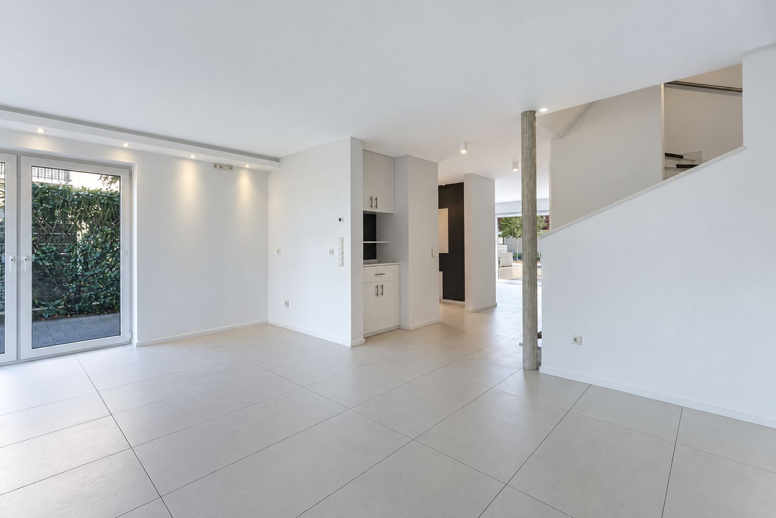 Helles, leerstehendes Wohnzimmer mit Fliesenboden, Glastür und Treppe.