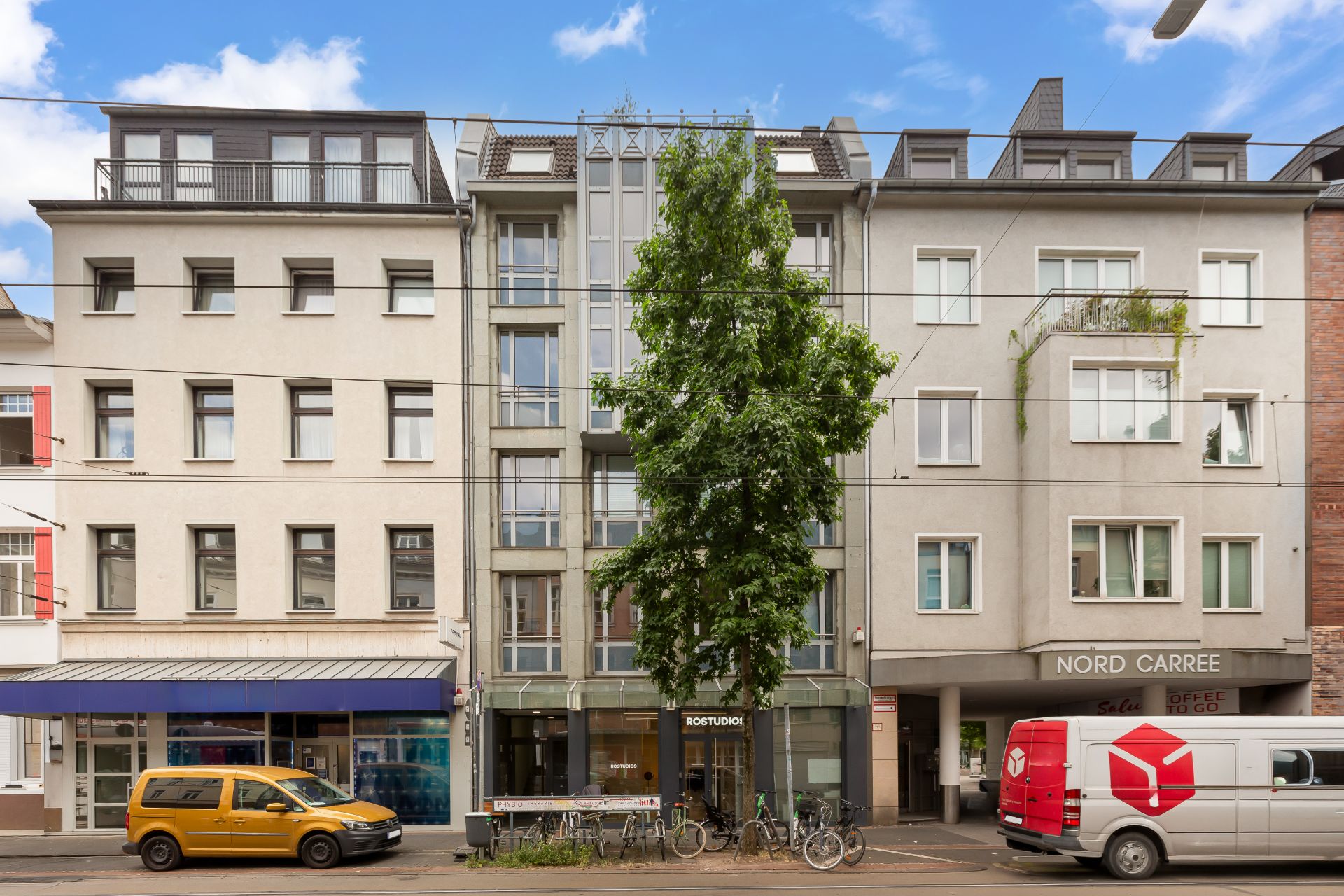 Mehrstöckiges Wohngebäude mit Balkonen, Bäumen und parkenden Autos an der Straße.