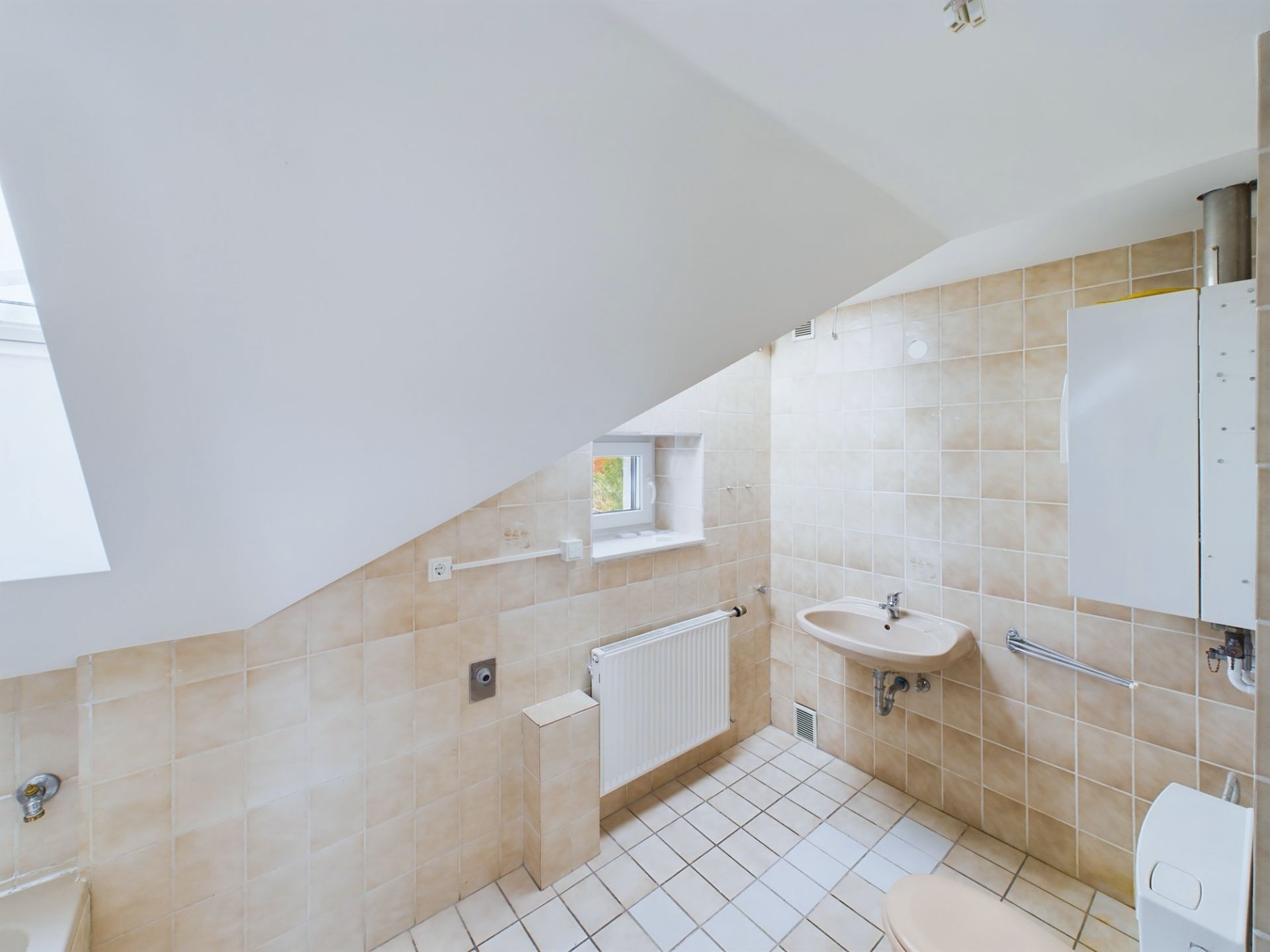 Helles Badezimmer mit beiger Fliesenwand, Waschbecken, kleinem Fenster und Heizkörper unter einer Dachschräge.