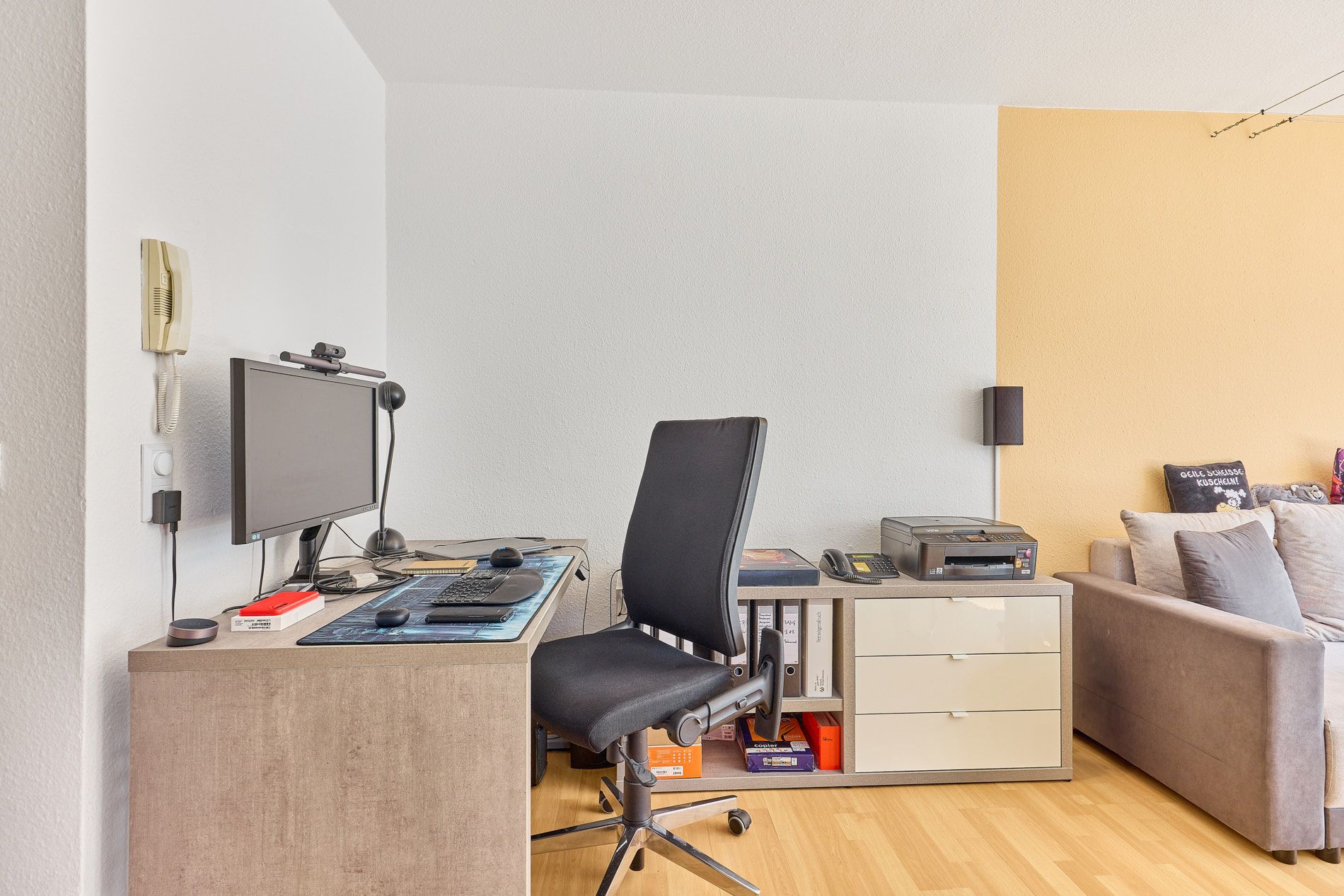 Arbeitsplatz mit Schreibtisch, Bürostuhl, Computer und Regalen neben einem grauen Sofa.