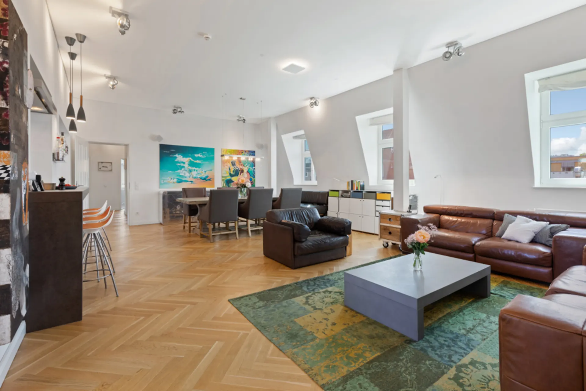 Modernes Wohnzimmer mit braunen Ledersofas, Esstisch, bunter Kunst und großen Fenstern.