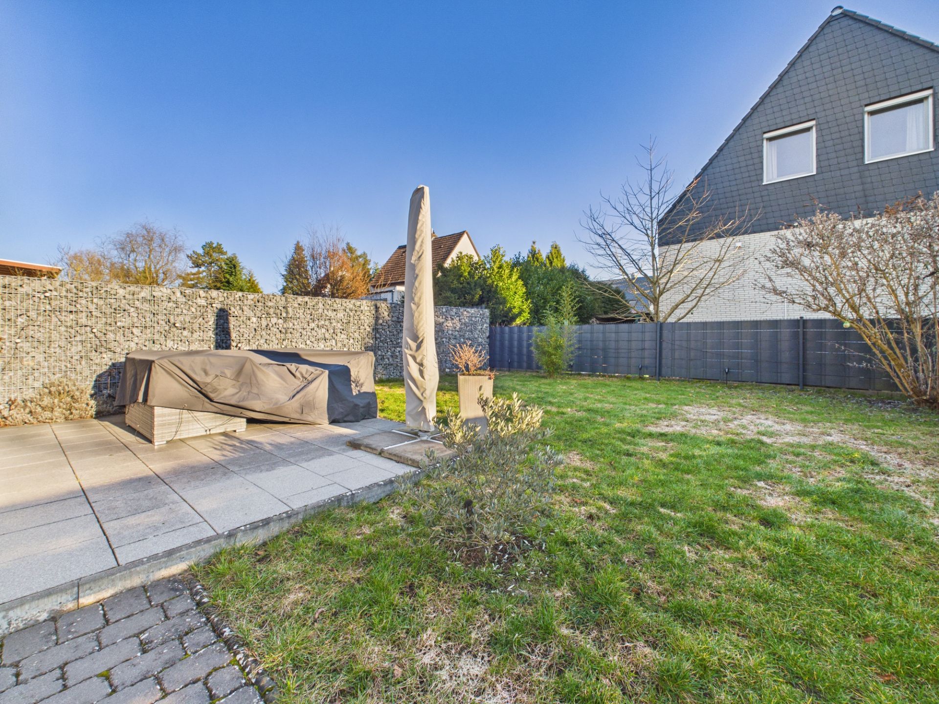 Garten mit Rasenfläche, Terrasse mit Abdeckung und geschlossener Sonnenschirm, angrenzende Steinumzäunung.