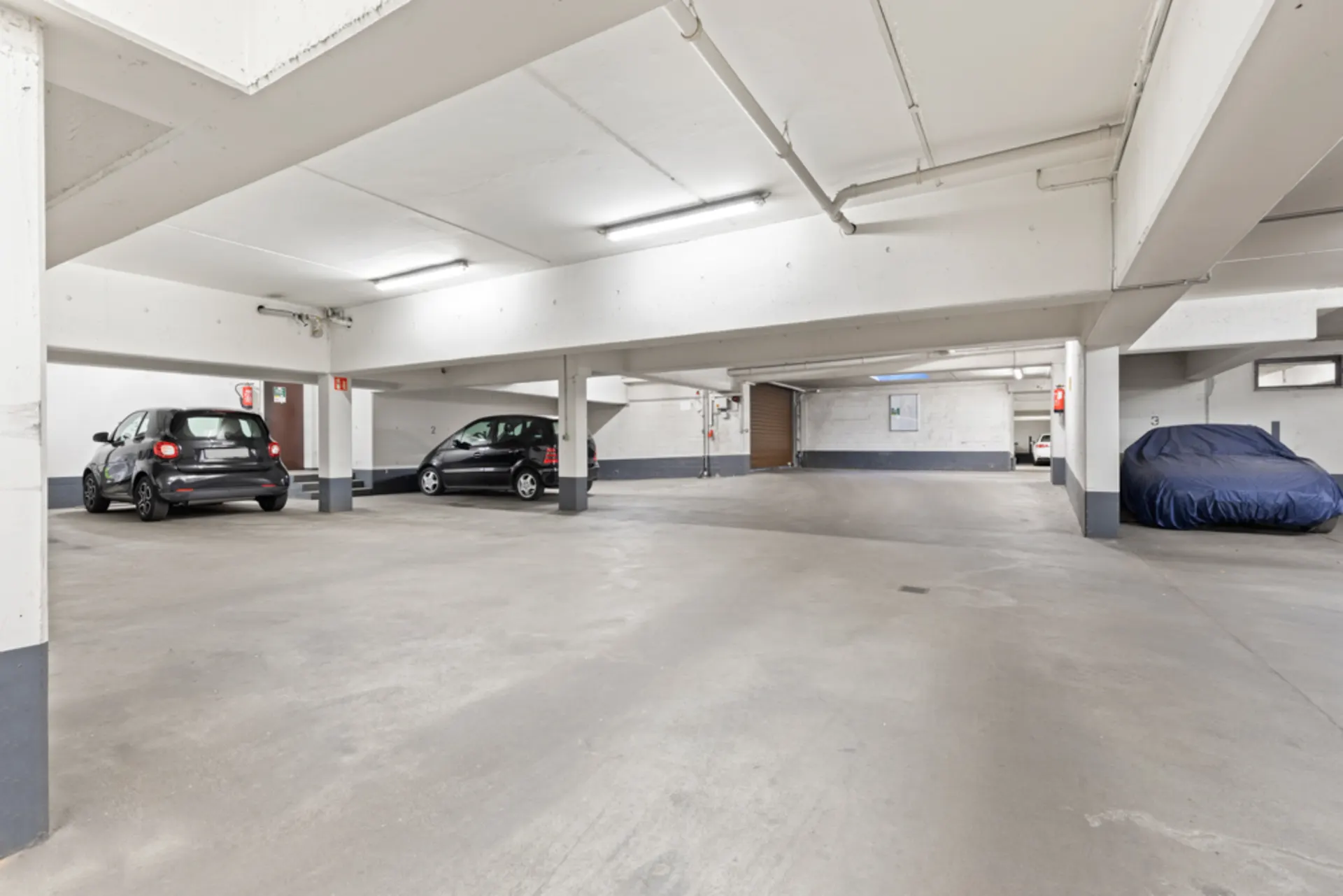 Helle Tiefgarage mit drei geparkten Autos, darunter ein Auto mit Abdeckung.
