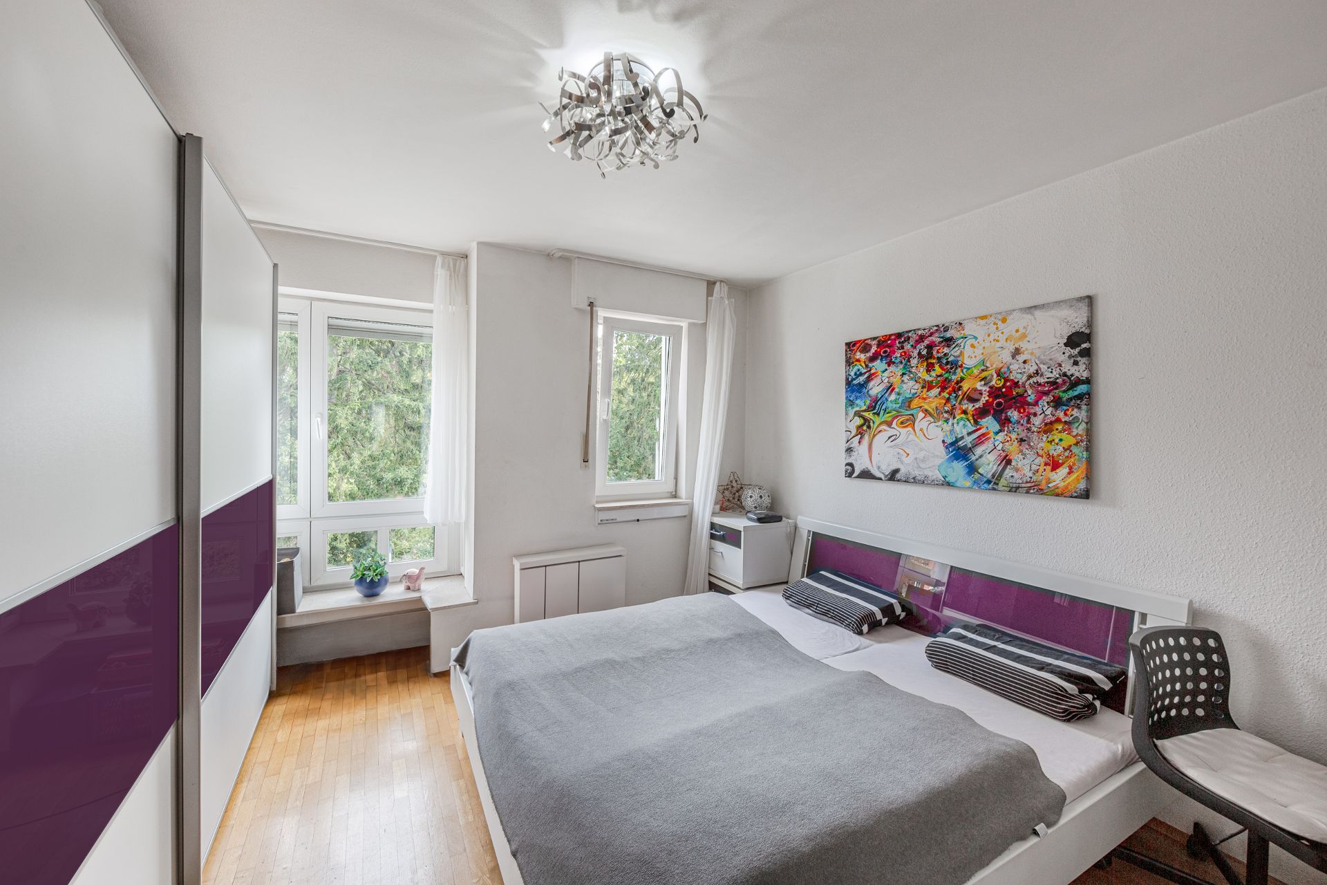 Helles Schlafzimmer mit Doppelbett, Kleiderschrank, buntem Bild und zwei Fenstern.