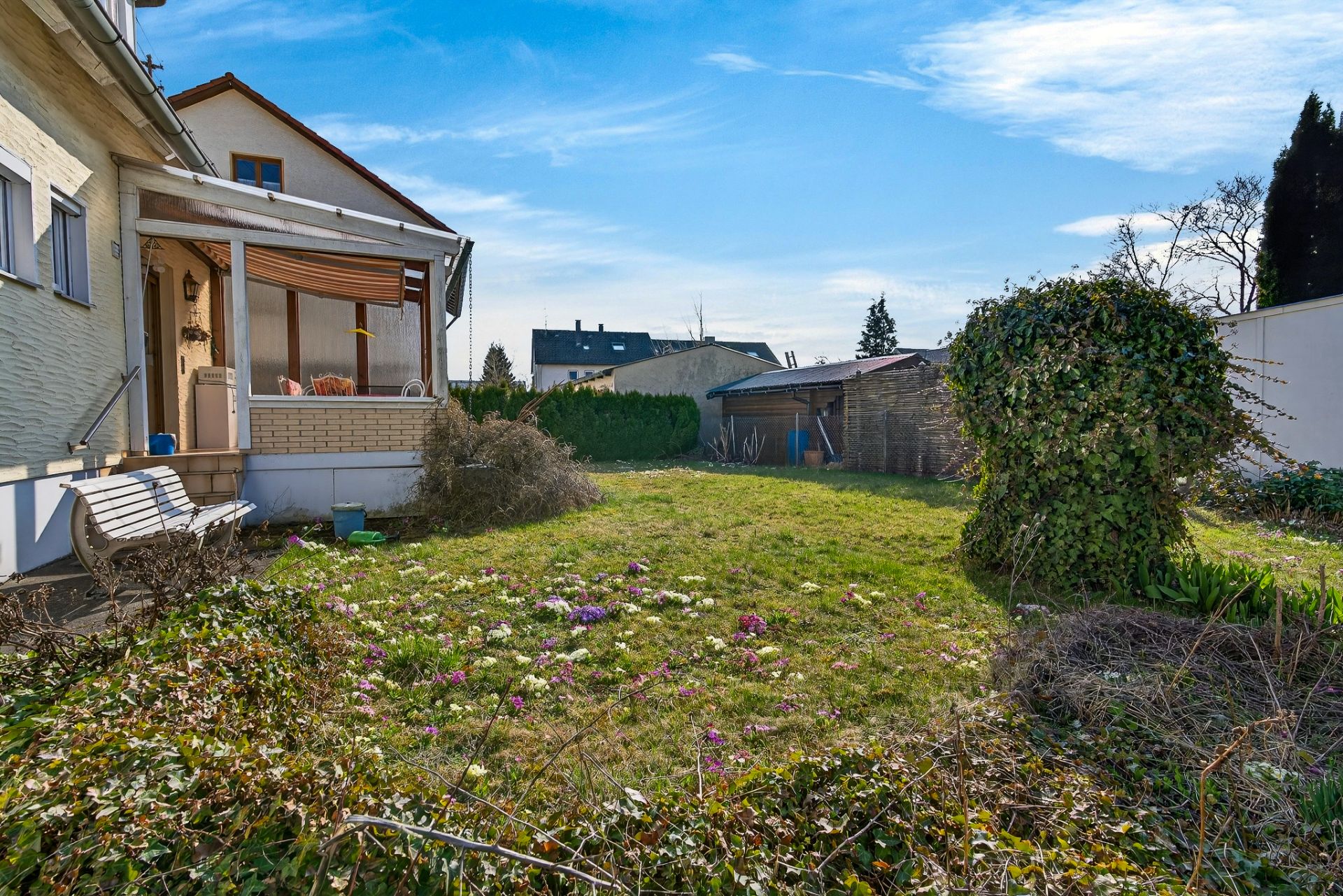 Garten mit Rasen, Blumen und Terrasse an einem Wohnhaus bei blauem Himmel.