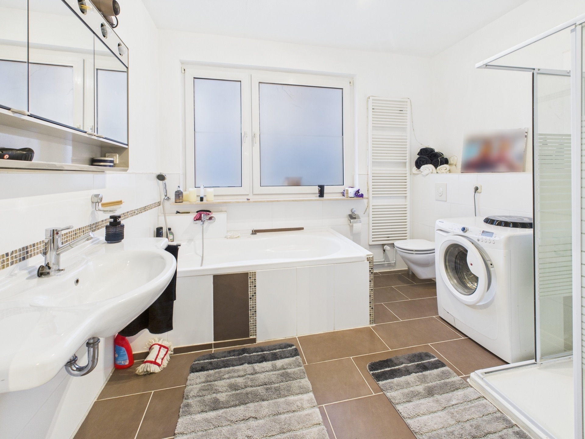 Helles Bad mit Badewanne, Waschbecken, WC, Waschmaschine und Dusche, zwei Fenster, braune Fliesen.