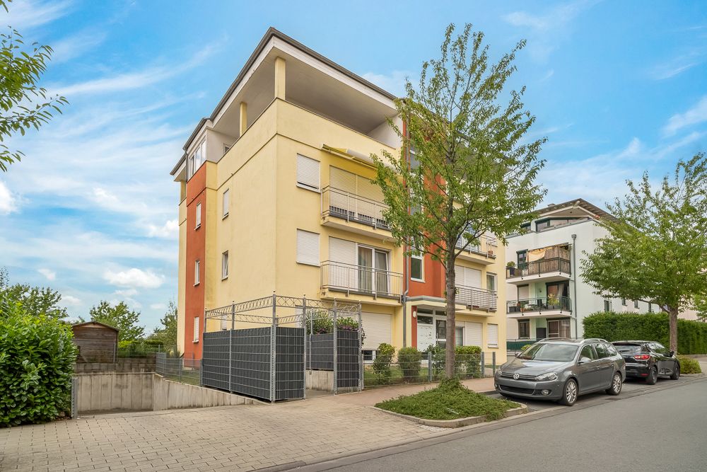 Mehrfamilienhaus mit Balkonen an einer ruhigen Straße, davor parken Autos, Bäume und Gehweg sichtbar.