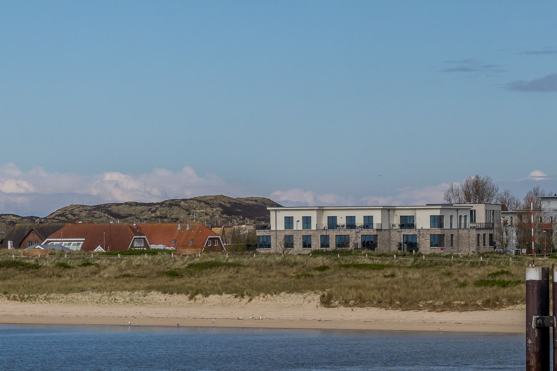 Wohnhäuser hinter einer Sanddüne mit Wasser im Vordergrund und Hügeln im Hintergrund.