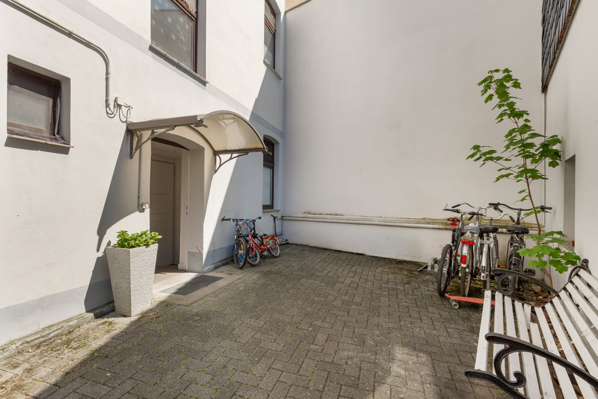 Gepflasterter Innenhof mit Eingang, Fahrrädern, Bank und kleinem Baum an einer weißen Hauswand.