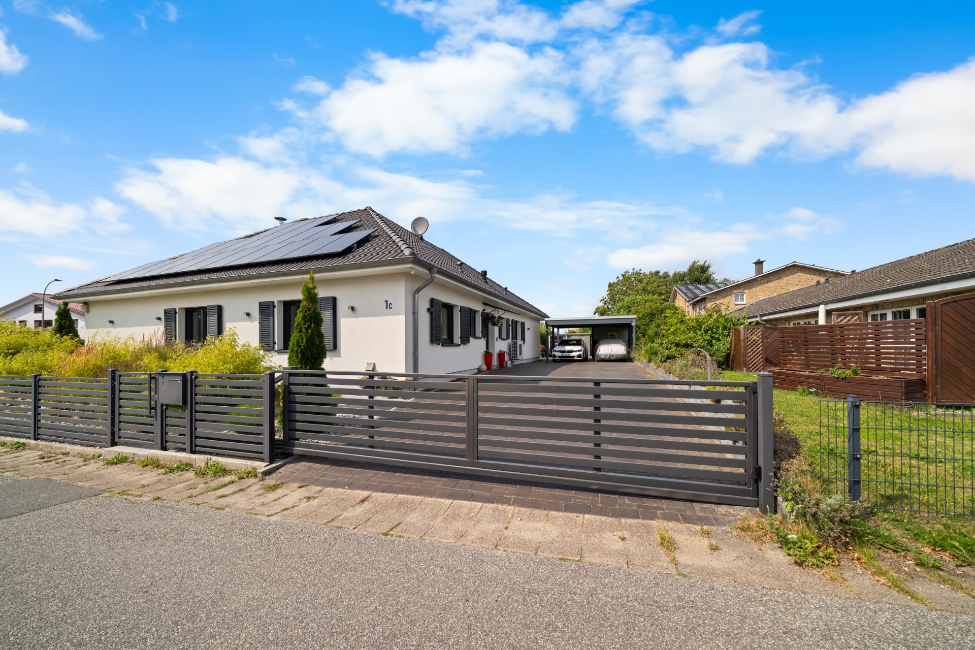 Modernes Einfamilienhaus mit Solarpanels, schwarzem Zaun und Carport bei Tag.