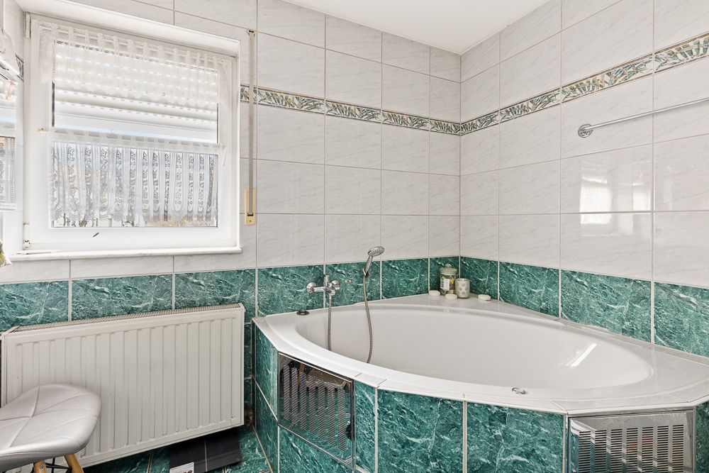 Eckbadewanne mit grün-weißen Fliesen, Fenster mit Rollos und Heizkörper daneben.