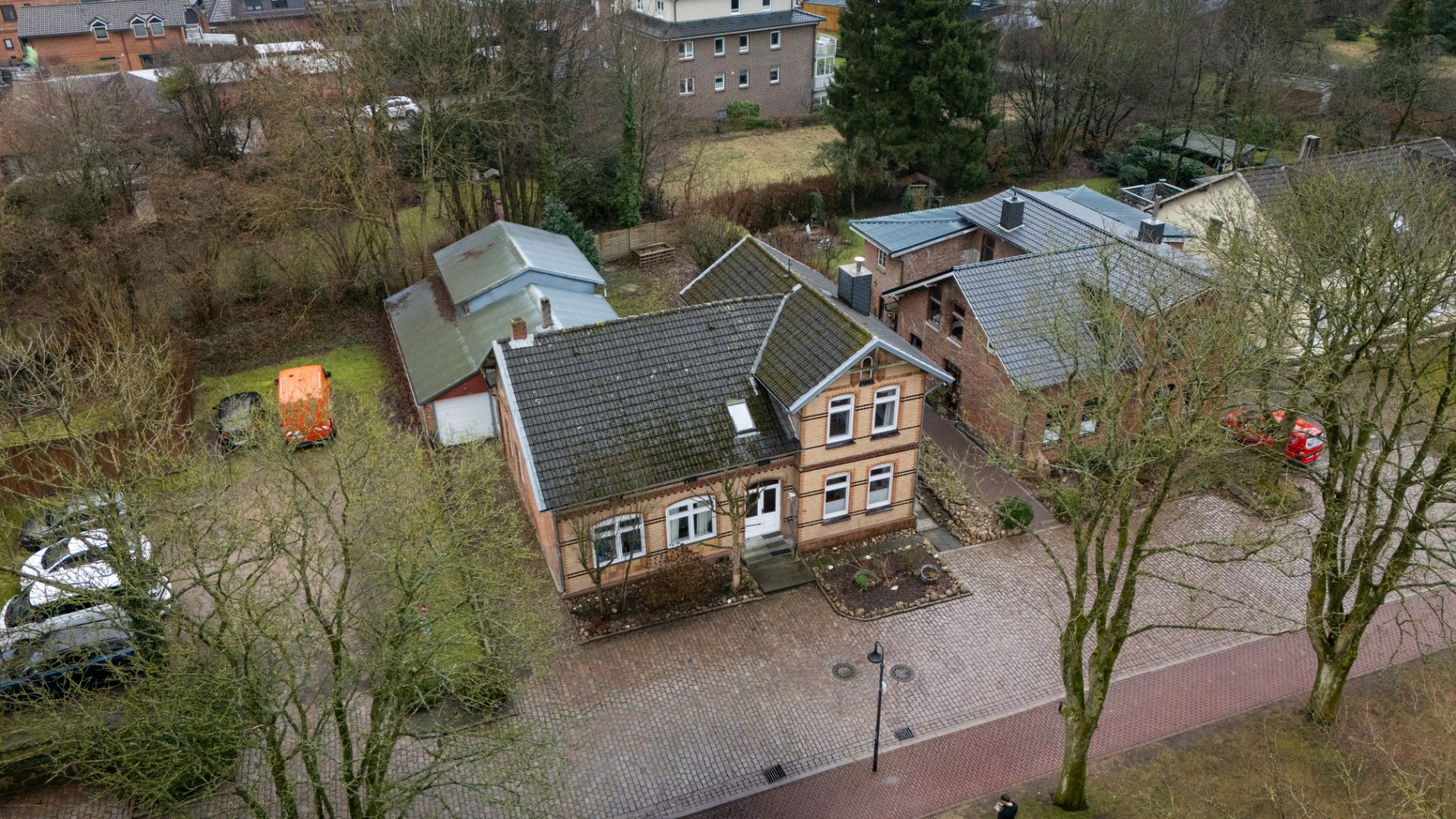 Backsteinhaus mit Satteldach und Vorhof, von Bäumen und Nachbarhäusern umgeben.