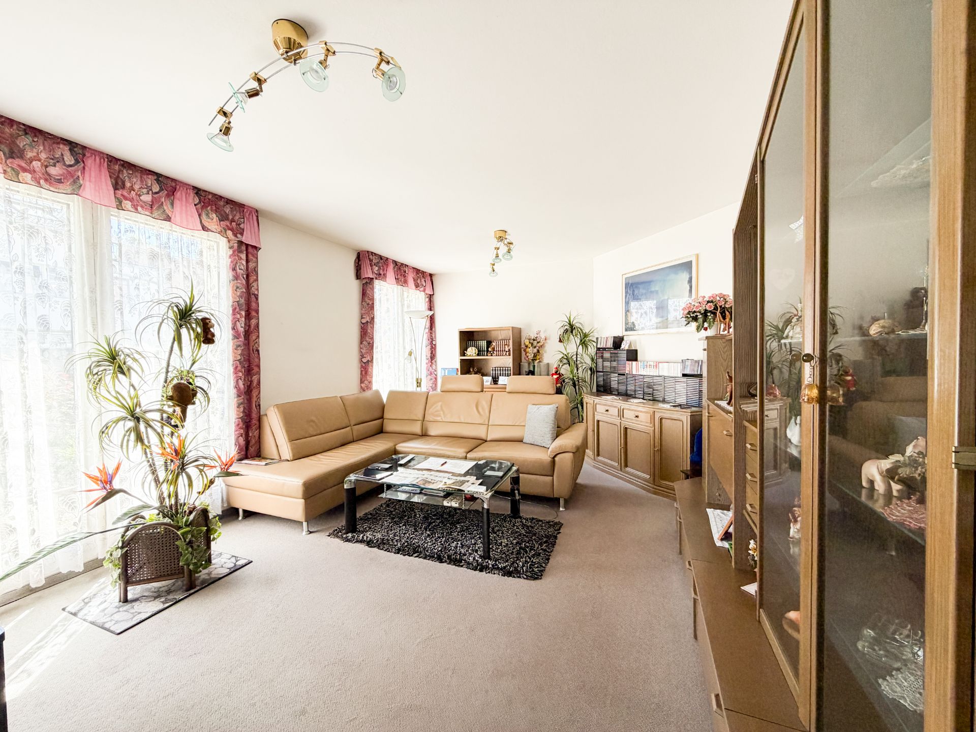 Helles Wohnzimmer mit Ecksofa, Couchtisch, Vitrine und großen Fenstern mit Vorhängen.