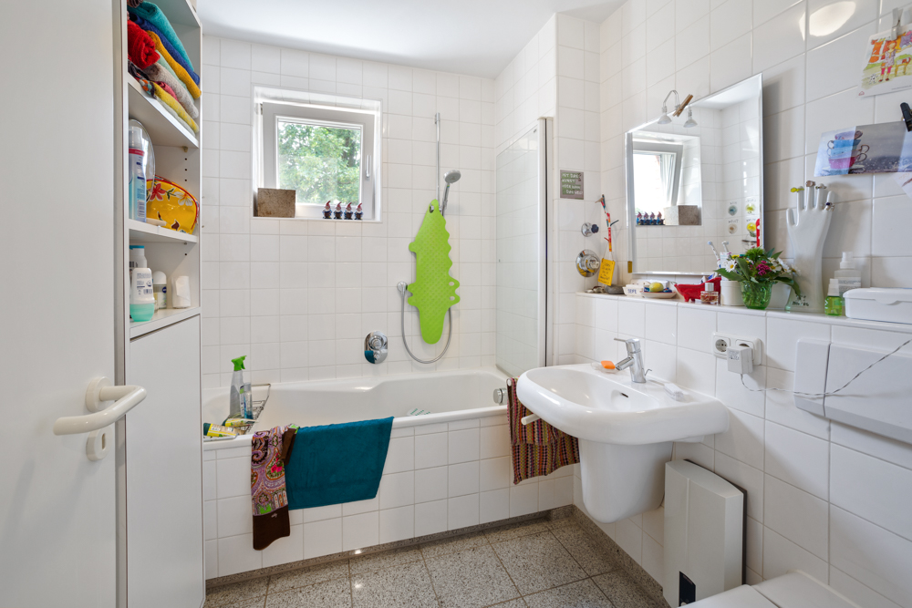 Helles Badezimmer mit Badewanne, Waschbecken, Regal und zwei Fenstern, weiße Fliesen an den Wänden.
