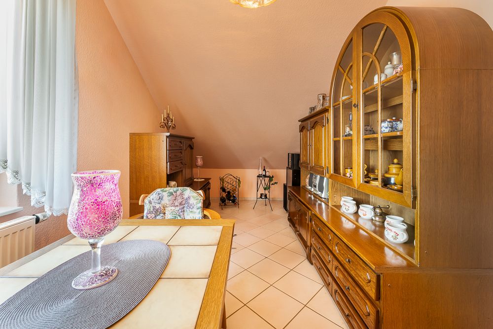 Wohnzimmer mit Holzschrank, Tisch, Stuhl und rosa Kerzenhalter, helle Fliesen und schräger Decke.