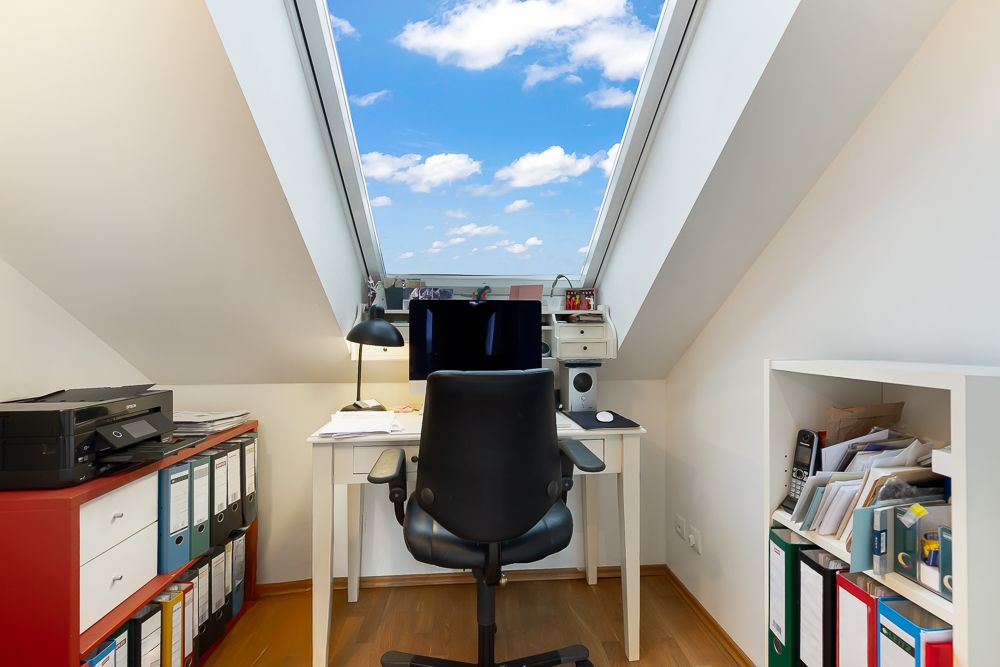 Arbeitszimmer unterm Dach mit Schreibtisch, Stuhl, Monitor und Regalen, Dachfenster mit Blick auf den Himmel.