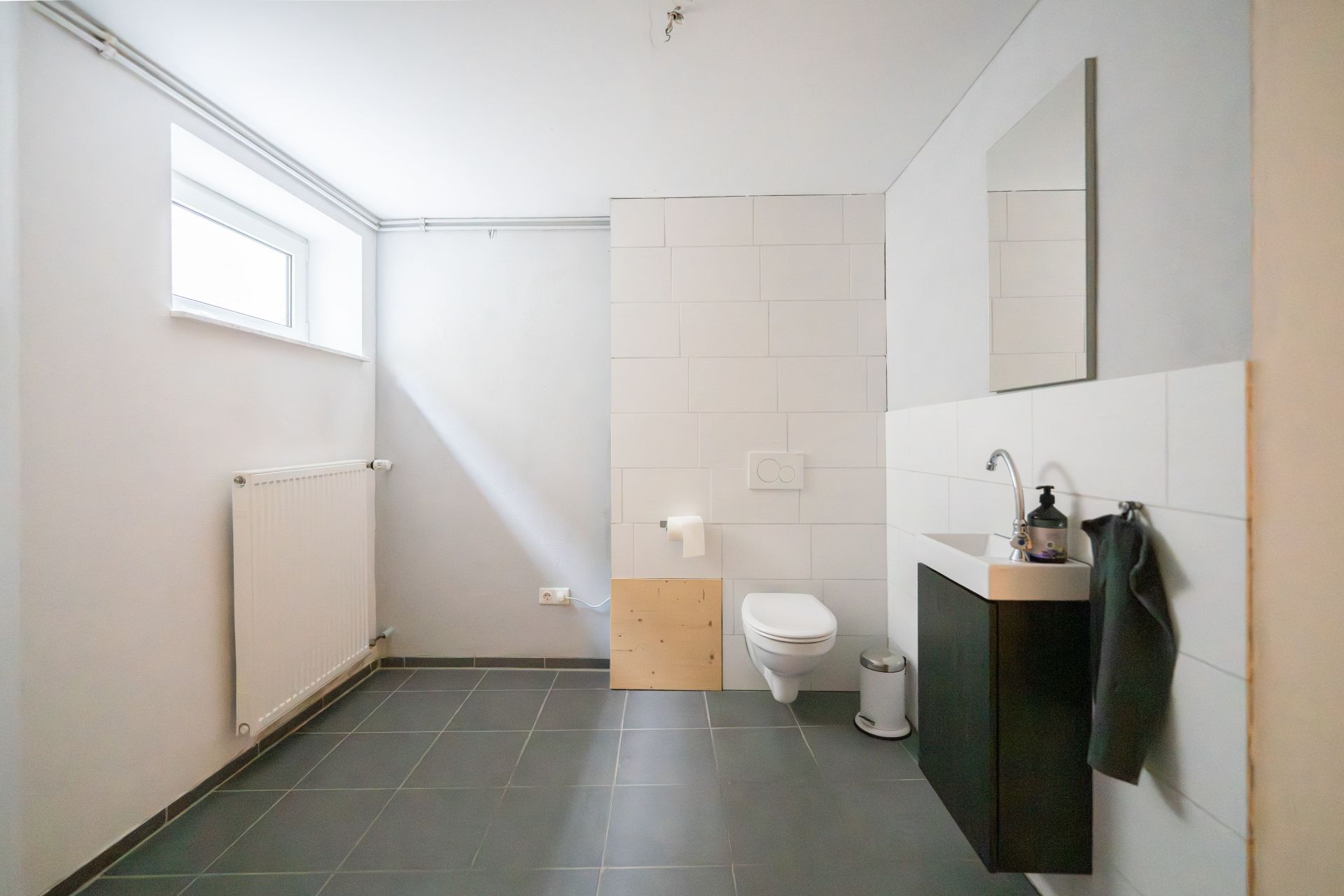Helles Badezimmer mit Wandtoilette, Waschbecken, Spiegel und Fenster.