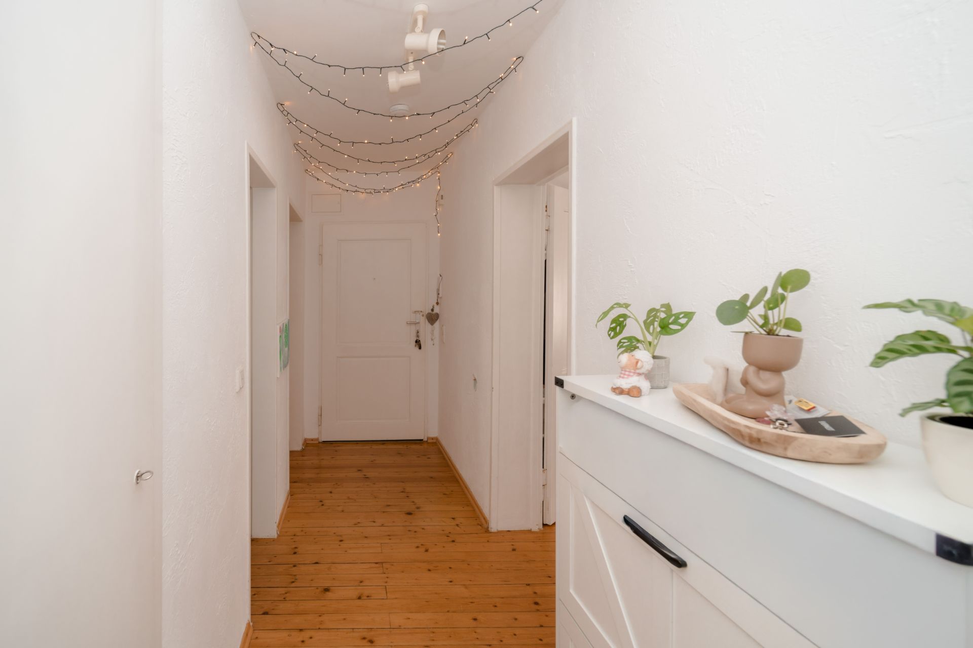 Heller Flur mit Holzboden, Dekolichter an der Decke und Pflanzen auf einem weißen Schrank.