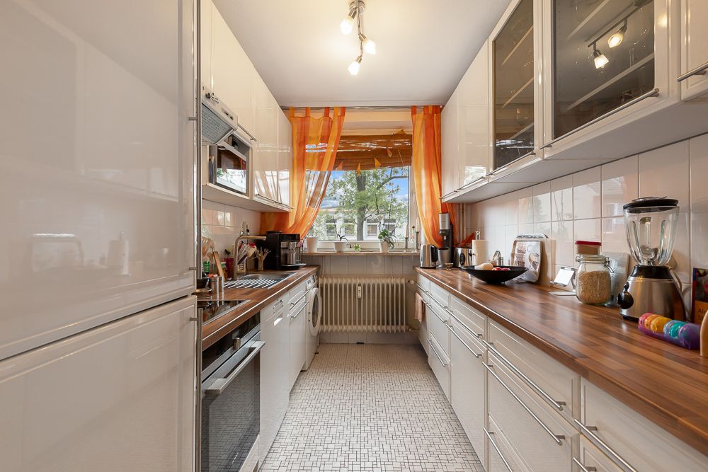 Schmale Küche mit weißen Schränken, Holzarbeitsplatten und einem Fenster mit orangefarbenen Vorhängen.