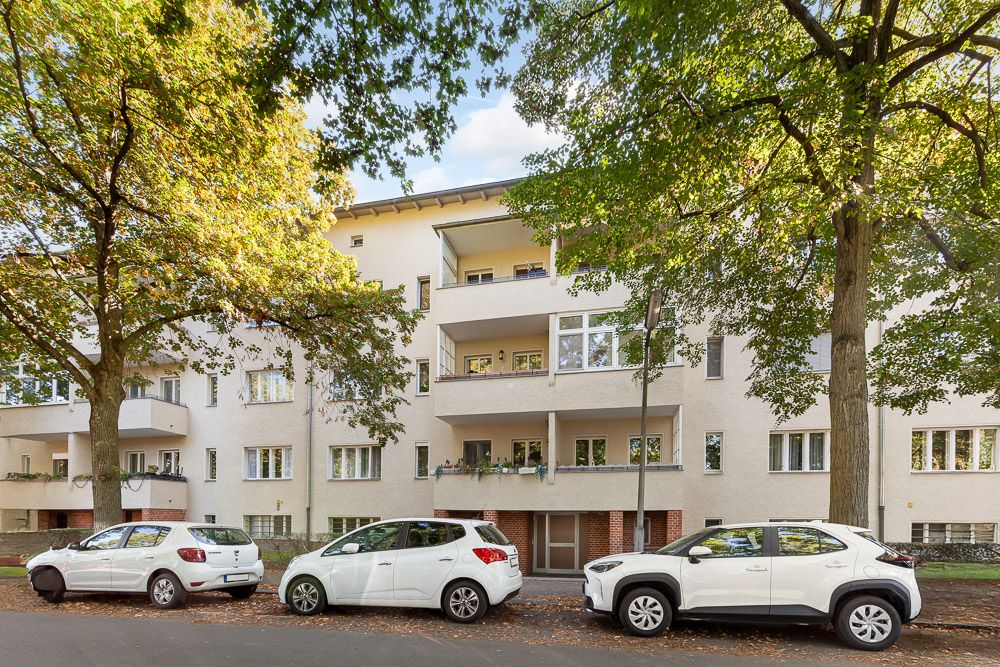 Mehrfamilienhaus mit Balkonen, davor geparkte Autos und große Bäume an der Straße.