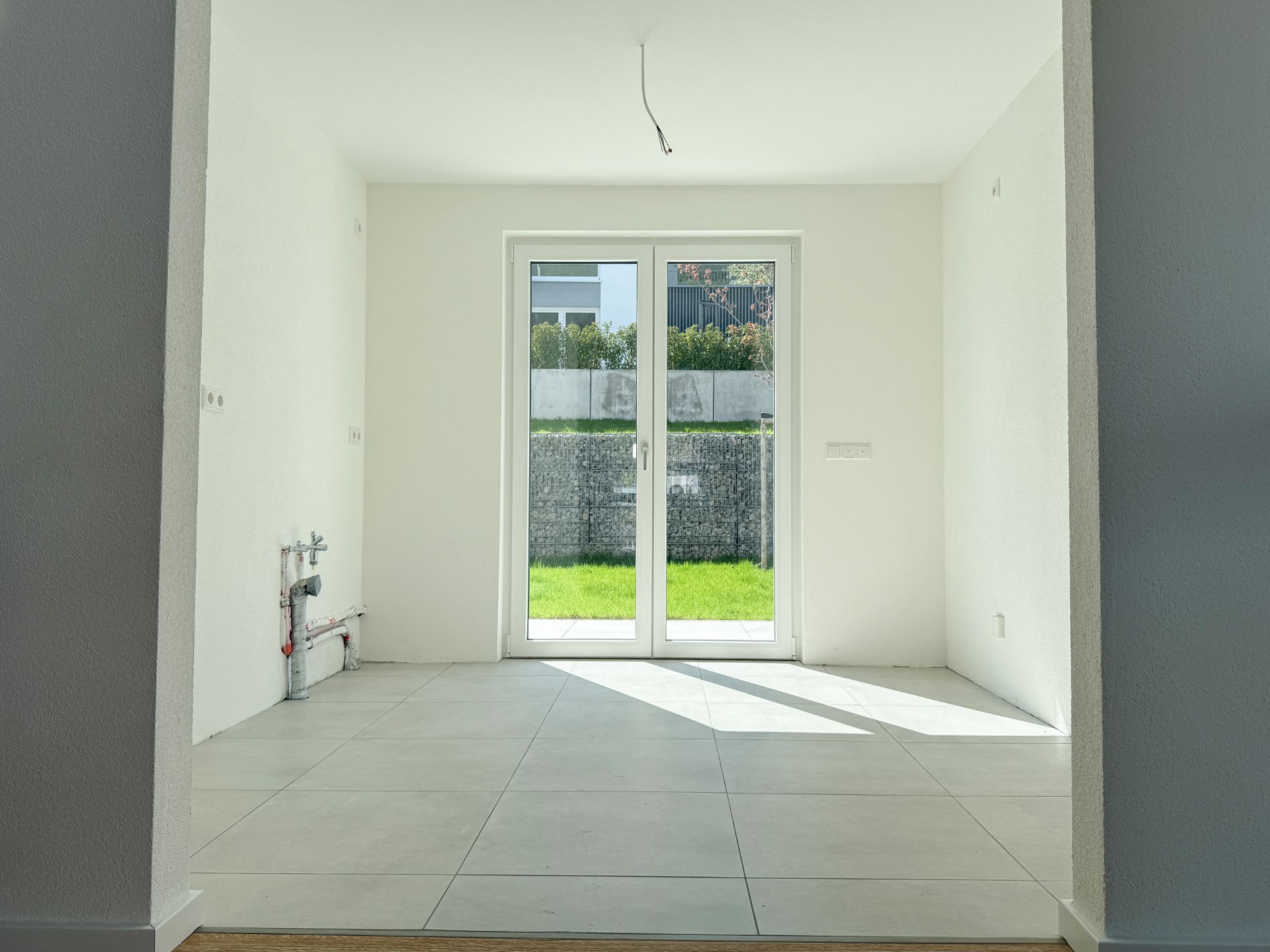 Heller leerer Raum mit Fliesenboden und großer Glastür zum Garten.