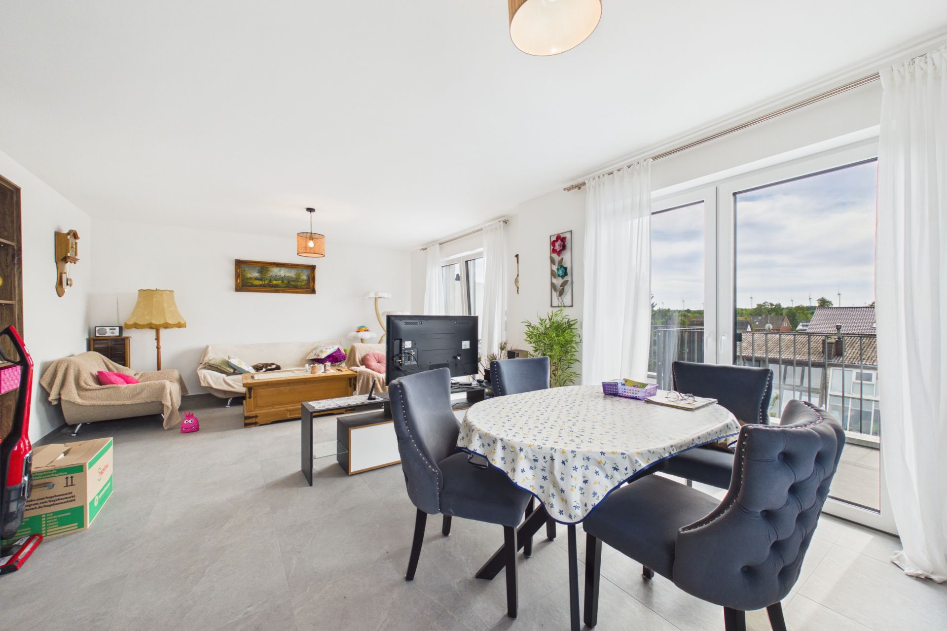 Helles Wohnzimmer mit Esstisch, Stühlen, Sofa, Fernseher und großen Fenstern.