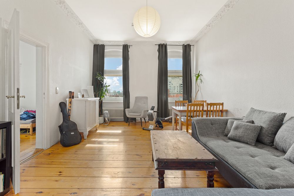 Helles Wohnzimmer mit Holzfußboden, grauem Sofa, Esstisch, Sessel und zwei Fenstern mit dunklen Vorhängen.