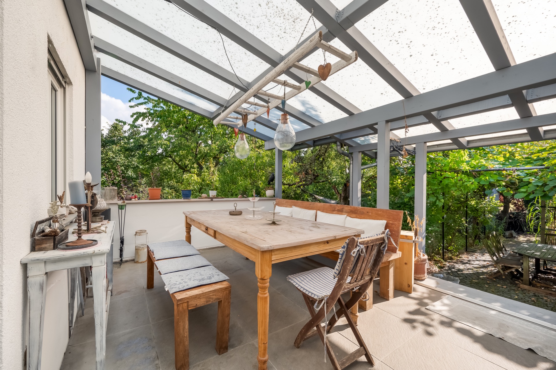 Überdachte Terrasse mit Holztisch, Bänken, Stühlen und Blick in einen grünen Garten.