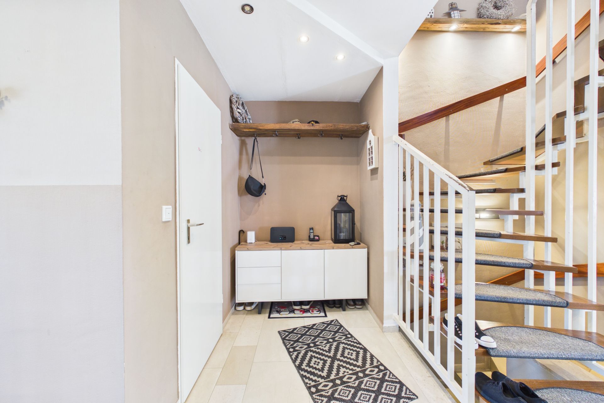Eingangsbereich mit weißer Tür, Kommode, Regal, Teppich und moderner Treppe mit Geländer.