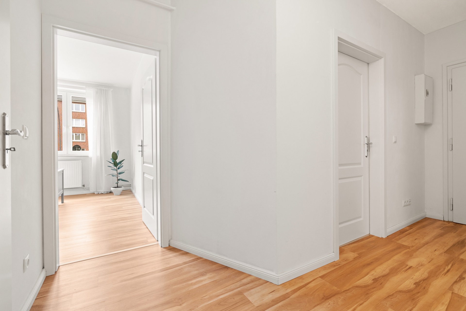 Heller Flur mit Holzboden, weißen Türen und Blick in ein Zimmer mit Fenster und Pflanze.