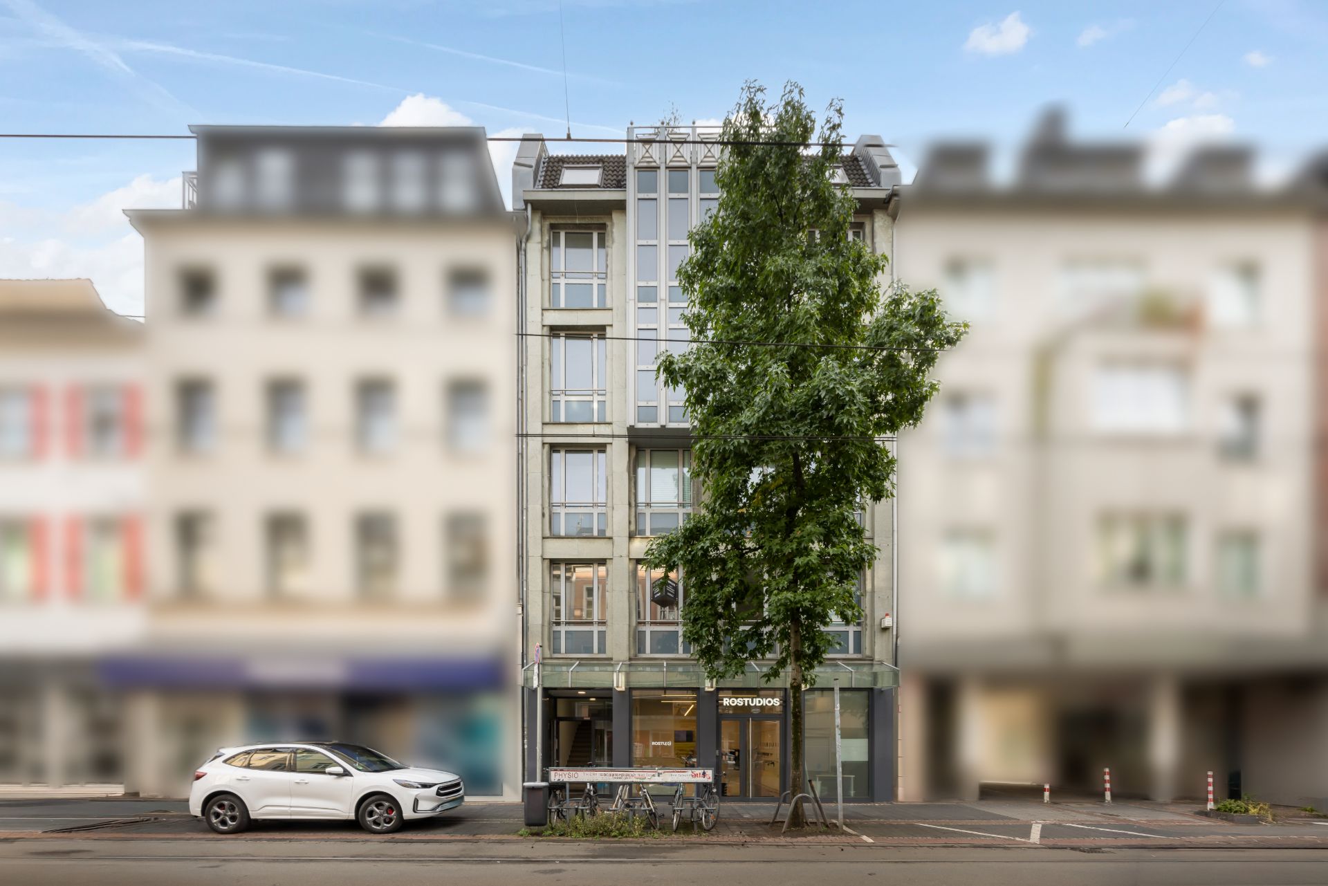 Mehrstöckiges Wohnhaus mit Glasfassade, Baum davor und weißem Auto auf der Straße.