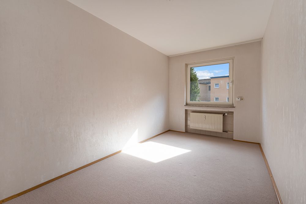 Leeres, helles Zimmer mit Teppichboden, Fenster und Heizkörper unter dem Fenster.