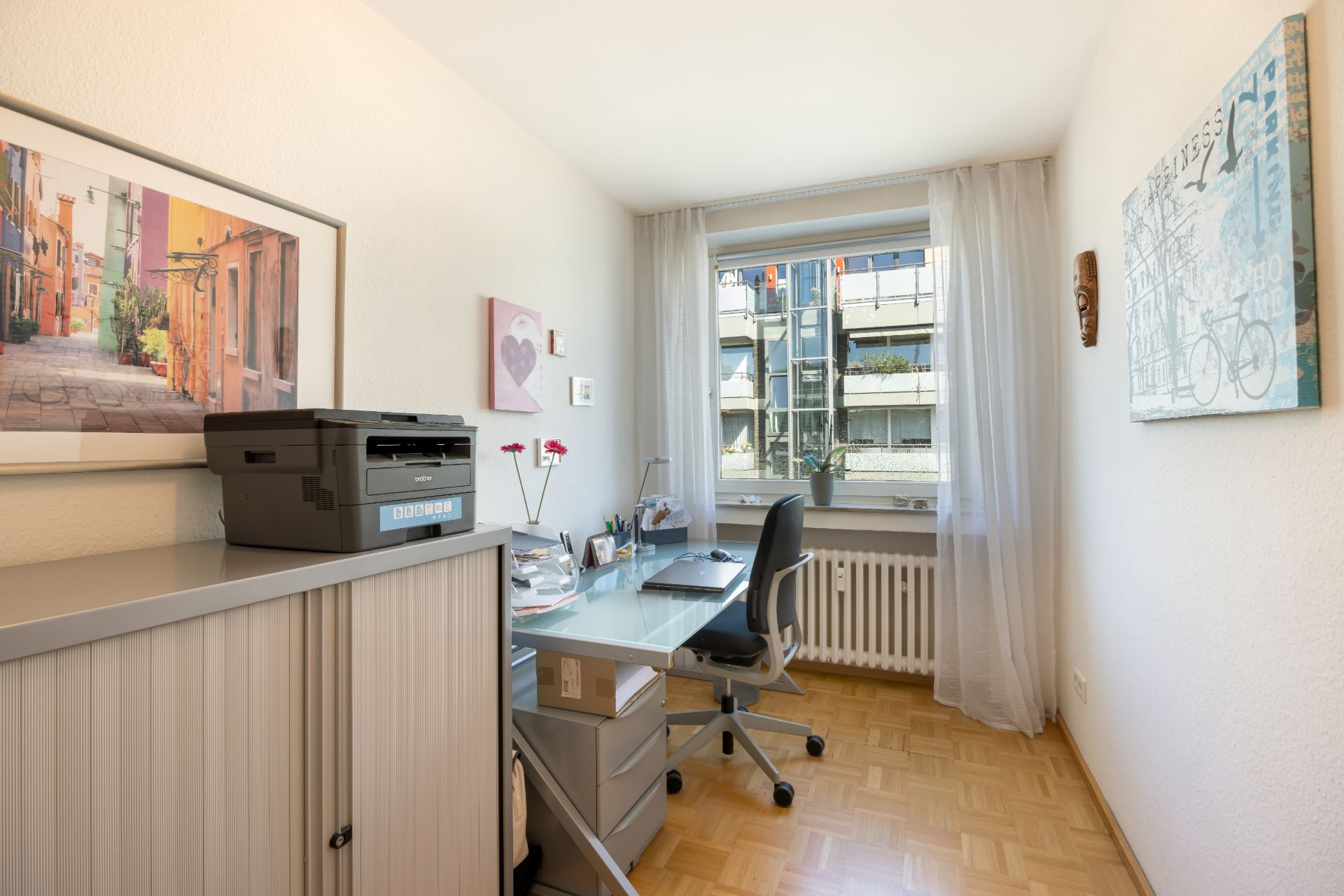 Helles Arbeitszimmer mit Schreibtisch, Stuhl, Drucker und Fenster mit Blick nach draußen.