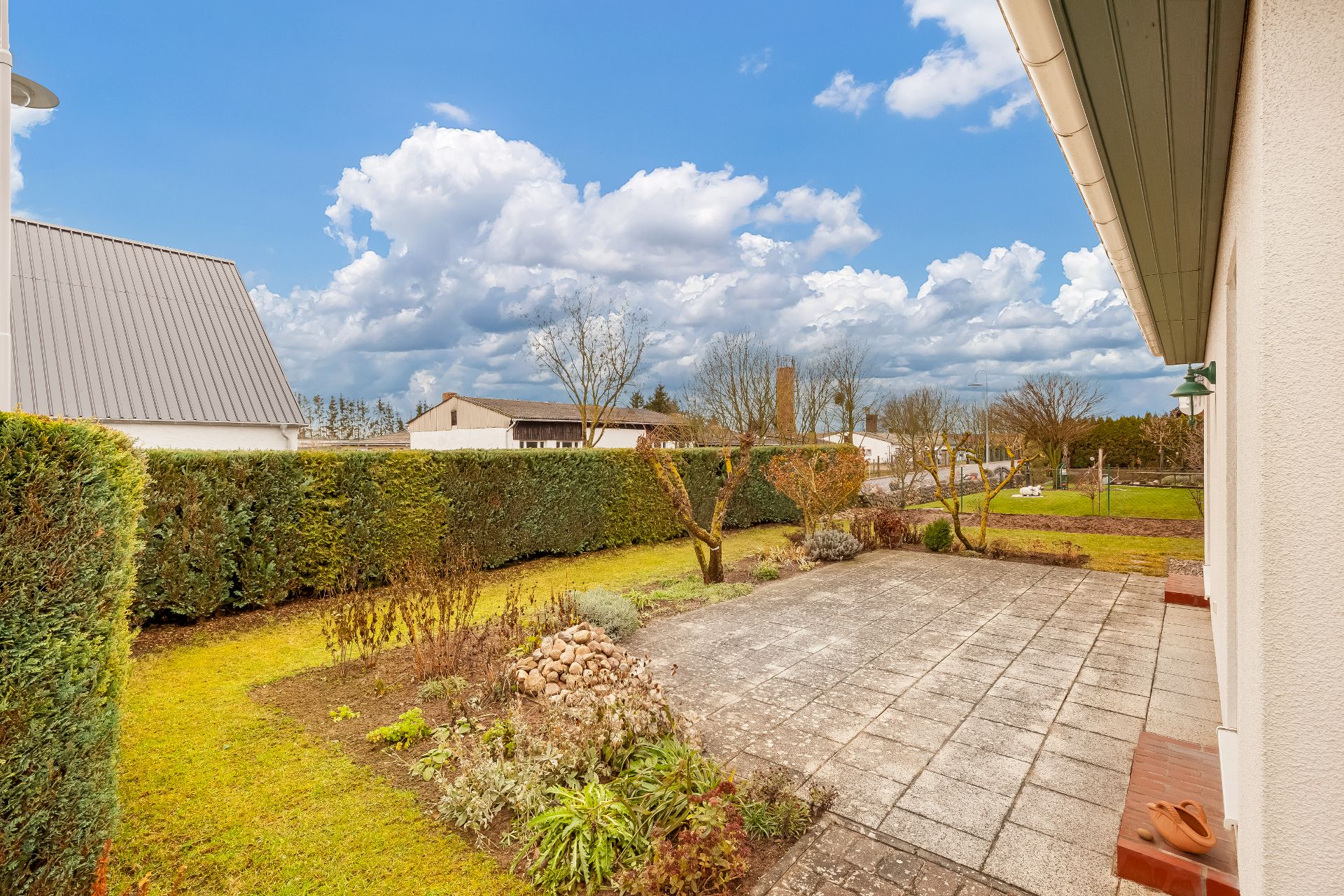 Gepflegter Garten mit Terrasse und Hecke, einige Bäume und Pflanzen, Hauswand rechts.