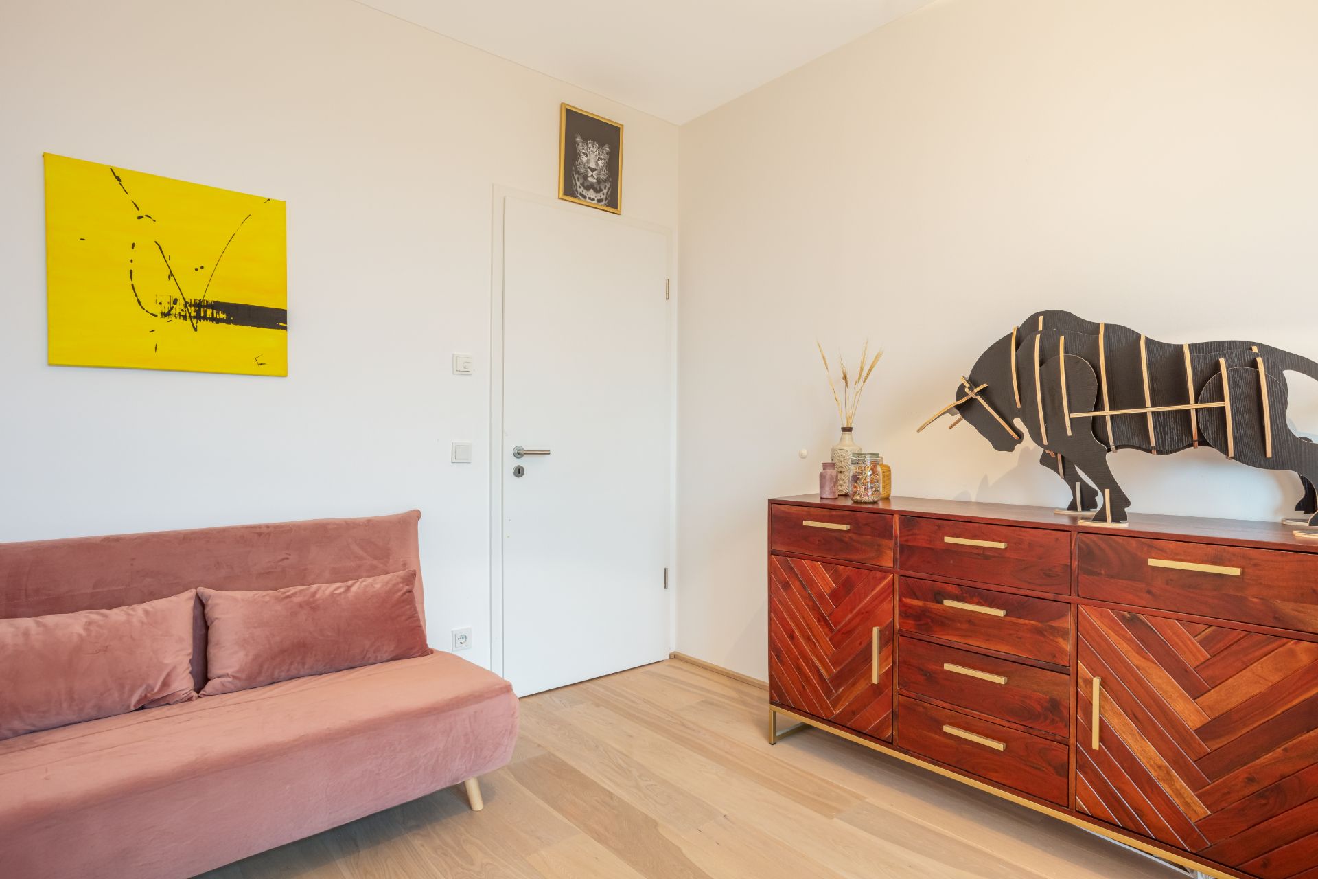 Zimmer mit rosa Sofa, Holzkommode, Dekorationsstier und gelbem Bild an der Wand.