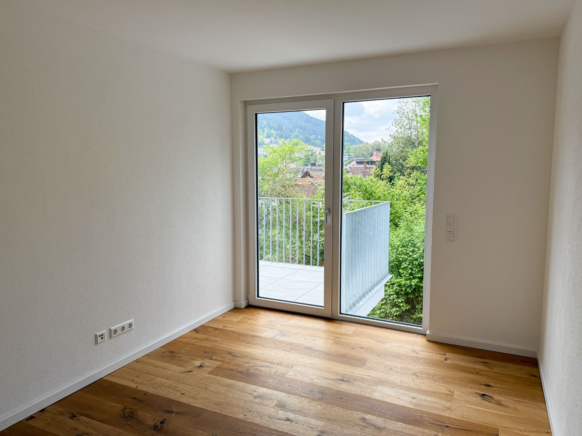 Leeres Zimmer mit Holzboden und großer Glastür zu einem Balkon mit Blick ins Grüne.