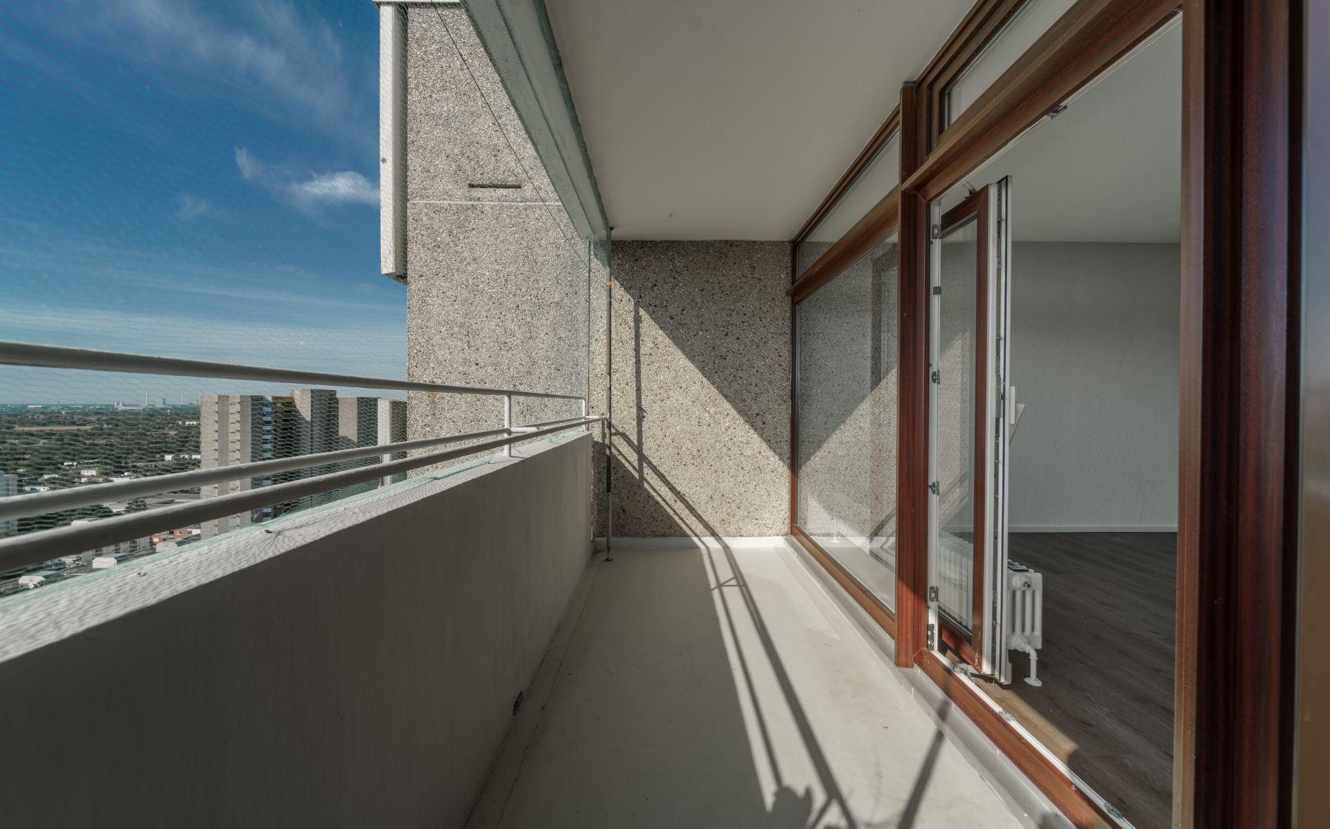 Balkon mit Betonwänden, Metallgeländer und Zugang zu einem Zimmer mit Holzboden.