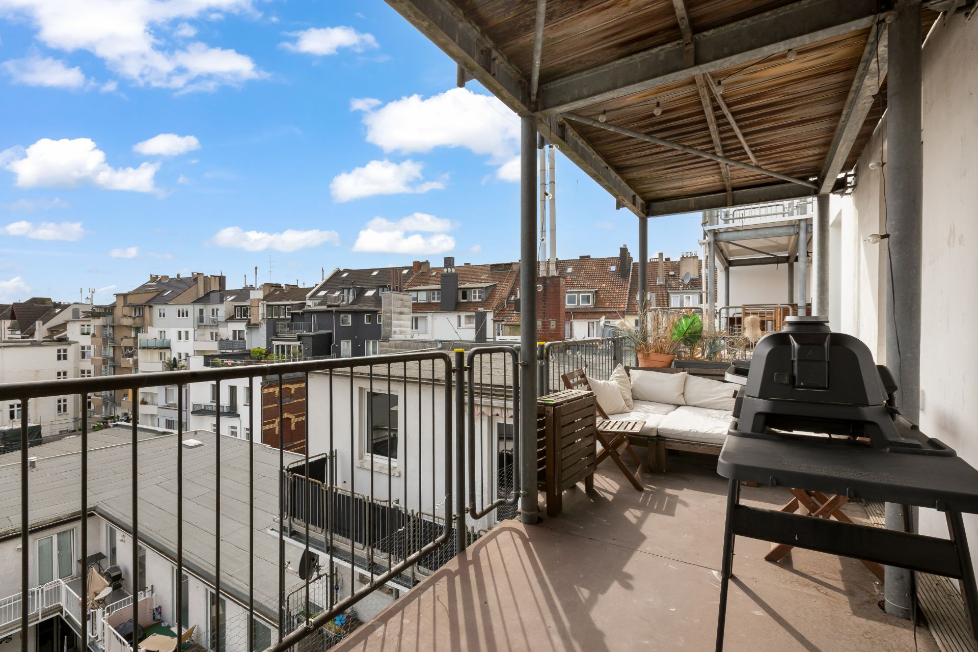 Überdachter Balkon mit Grill, Sitzbank und Blick auf umliegende Wohnhäuser.