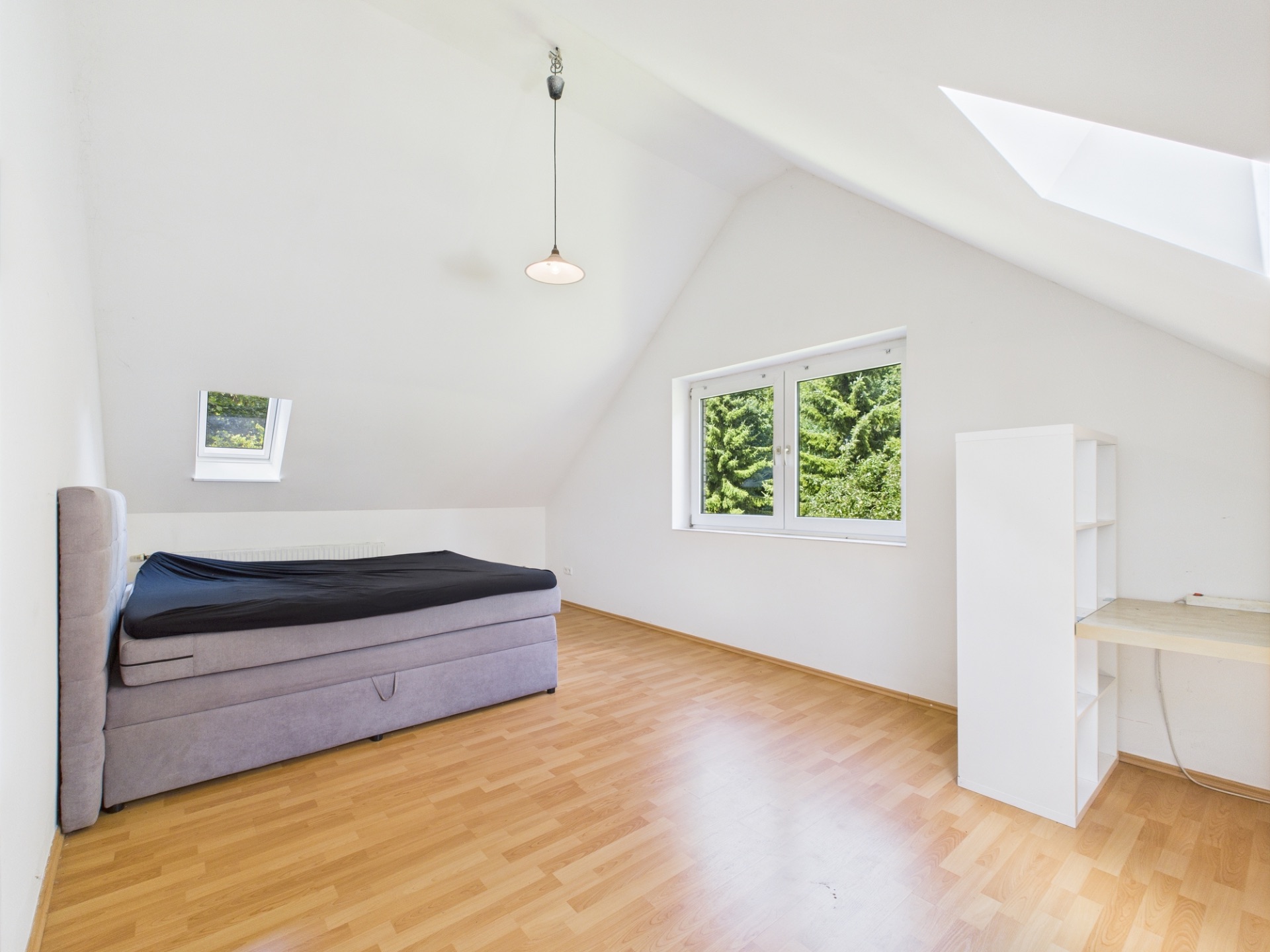 Helles Schlafzimmer mit Dachschräge, Bett, Schreibtisch und großem Fenster mit Blick ins Grüne.